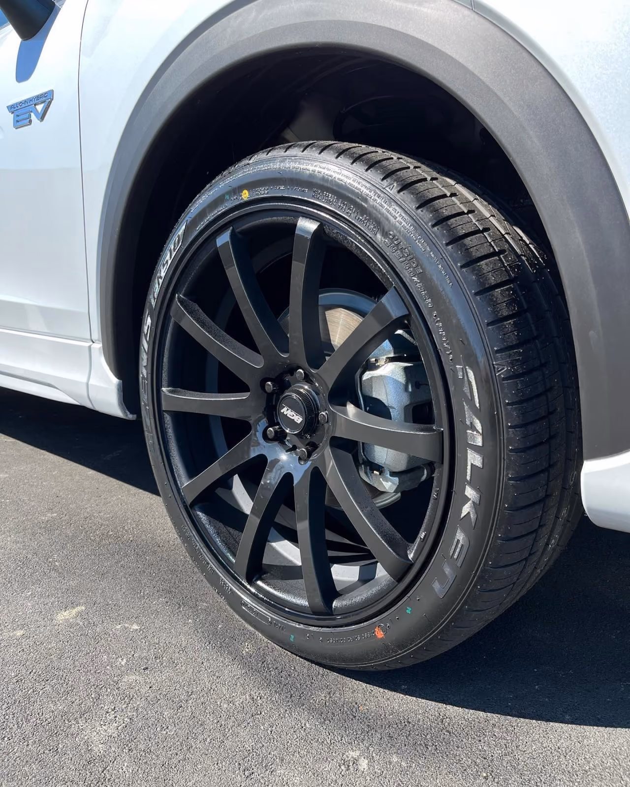 Close-up of a white plug-in hybrid electric vehicle's black alloy wheel and Falken tire on asphalt.