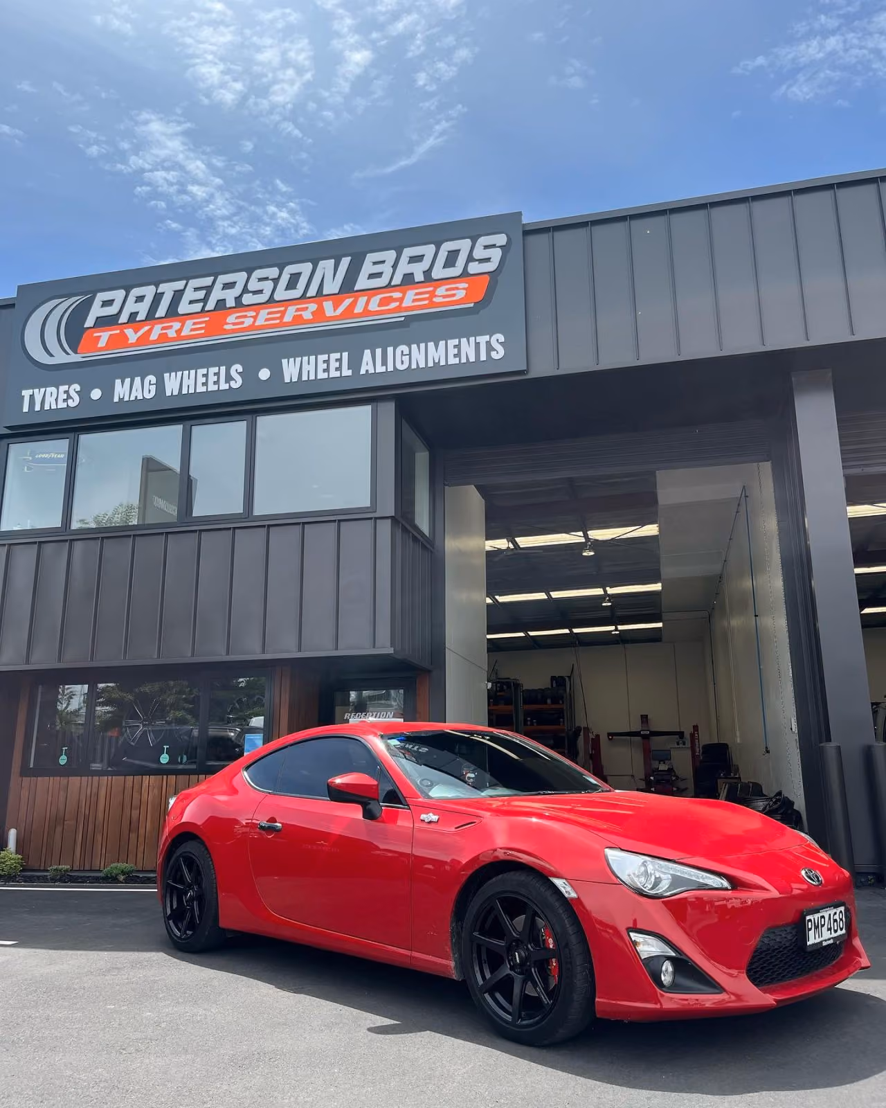 Red sports car with black wheels parked in front of Paterson Bros Tyre Services building.