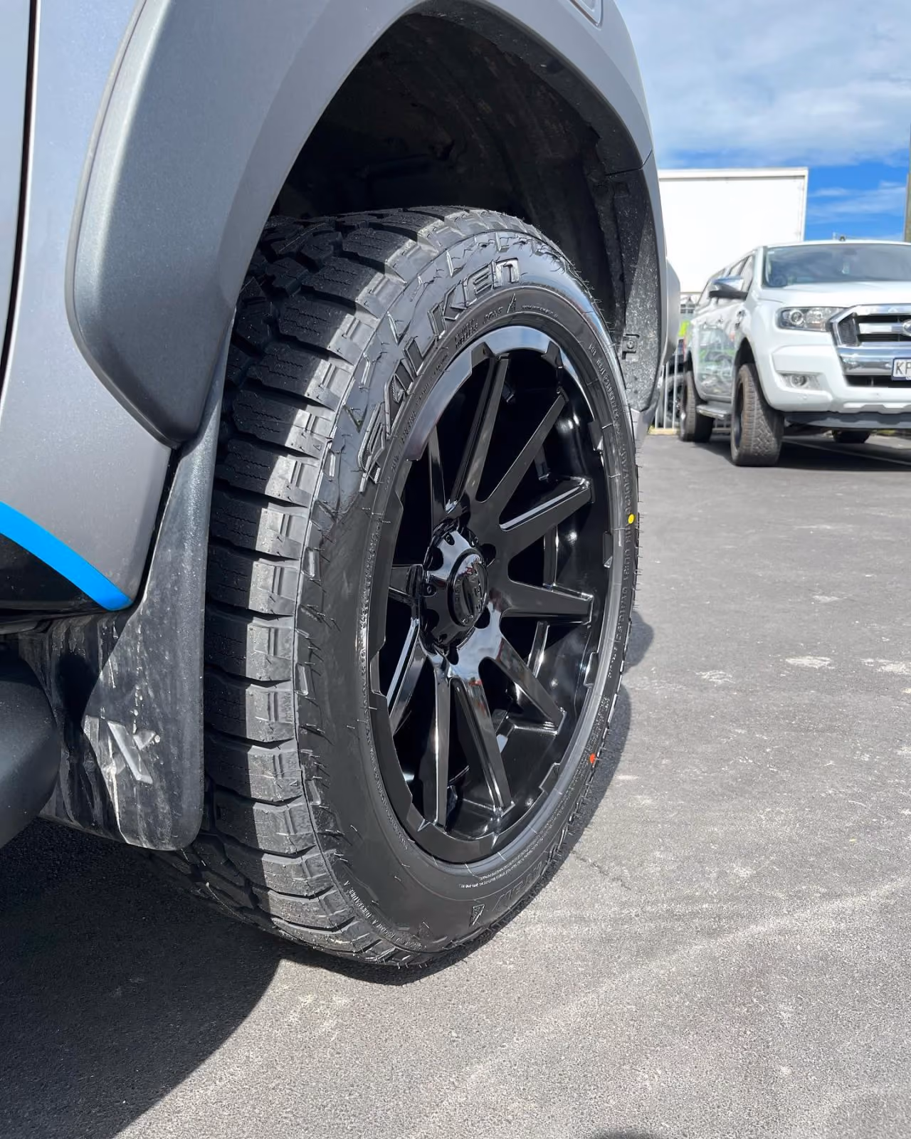 Close-up of a black Falken off-road tire on a silver truck with a white truck in the background.