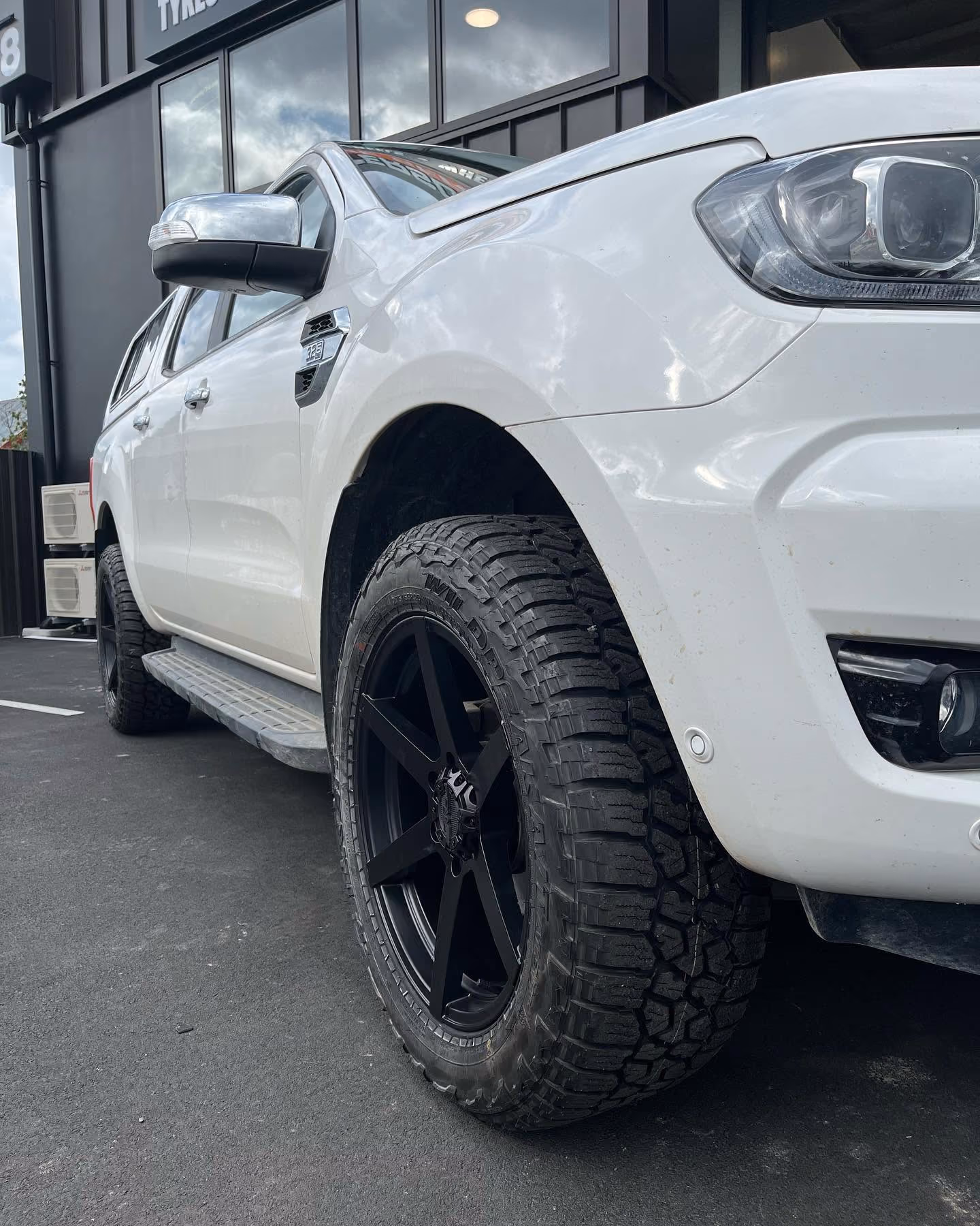 Close-up of a white pickup truck's front side, showing large black off-road tire and headlight.