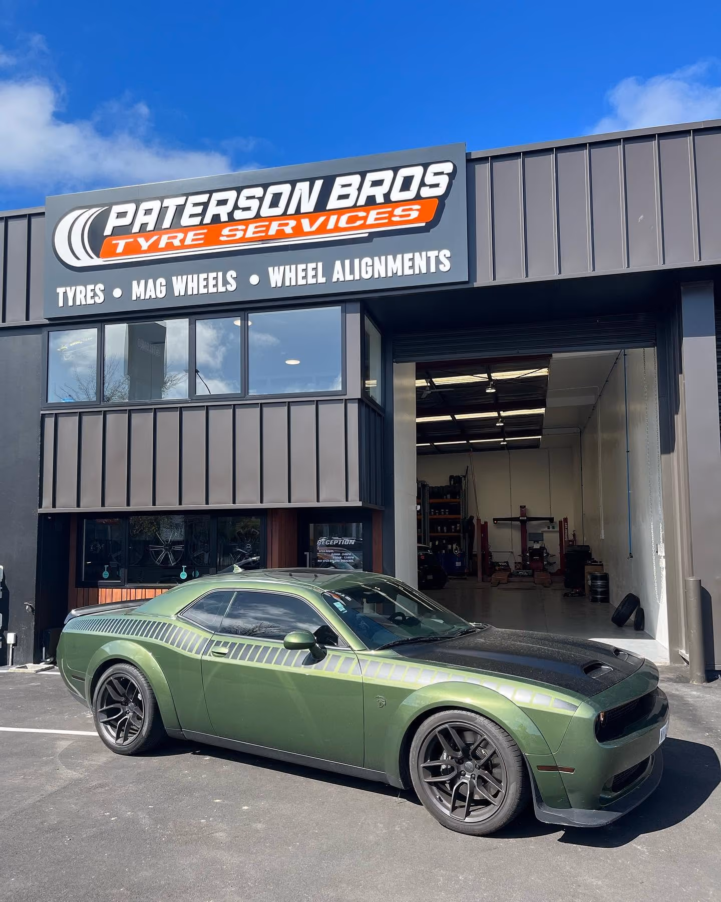 Green muscle car with black wheels parked outside Paterson Bros Tyre Services shop.