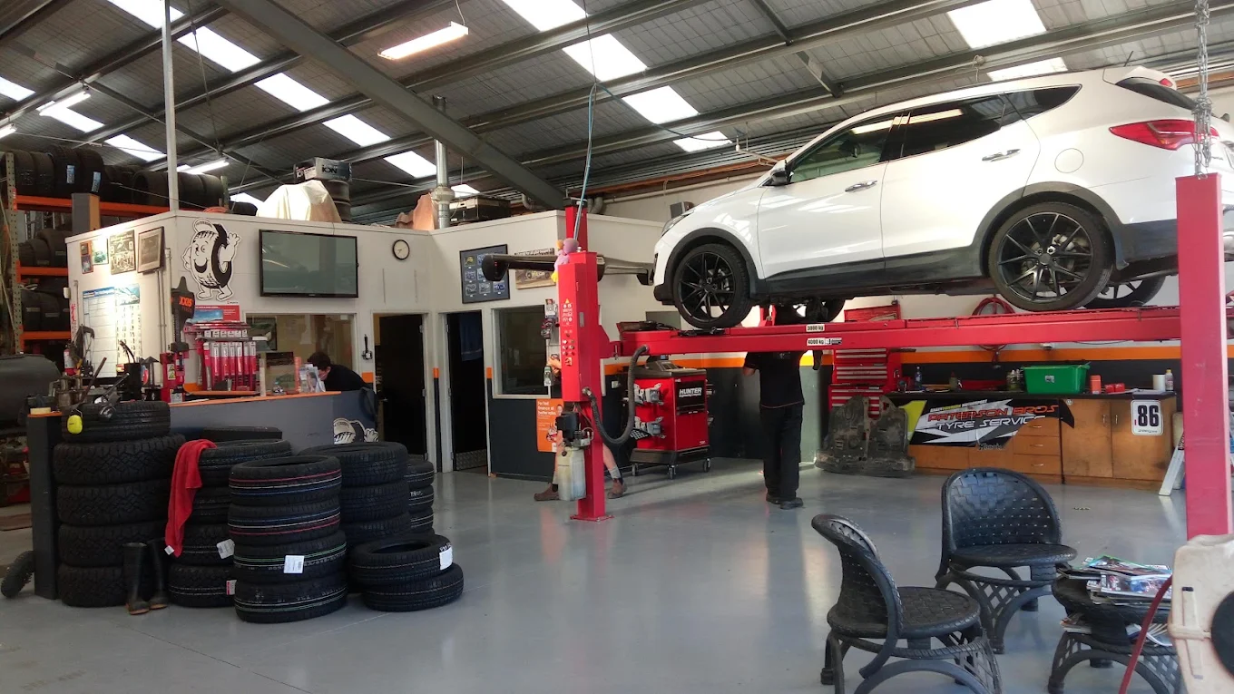 White SUV on a red hydraulic lift inside a tire service garage with stacked tires and chairs.
