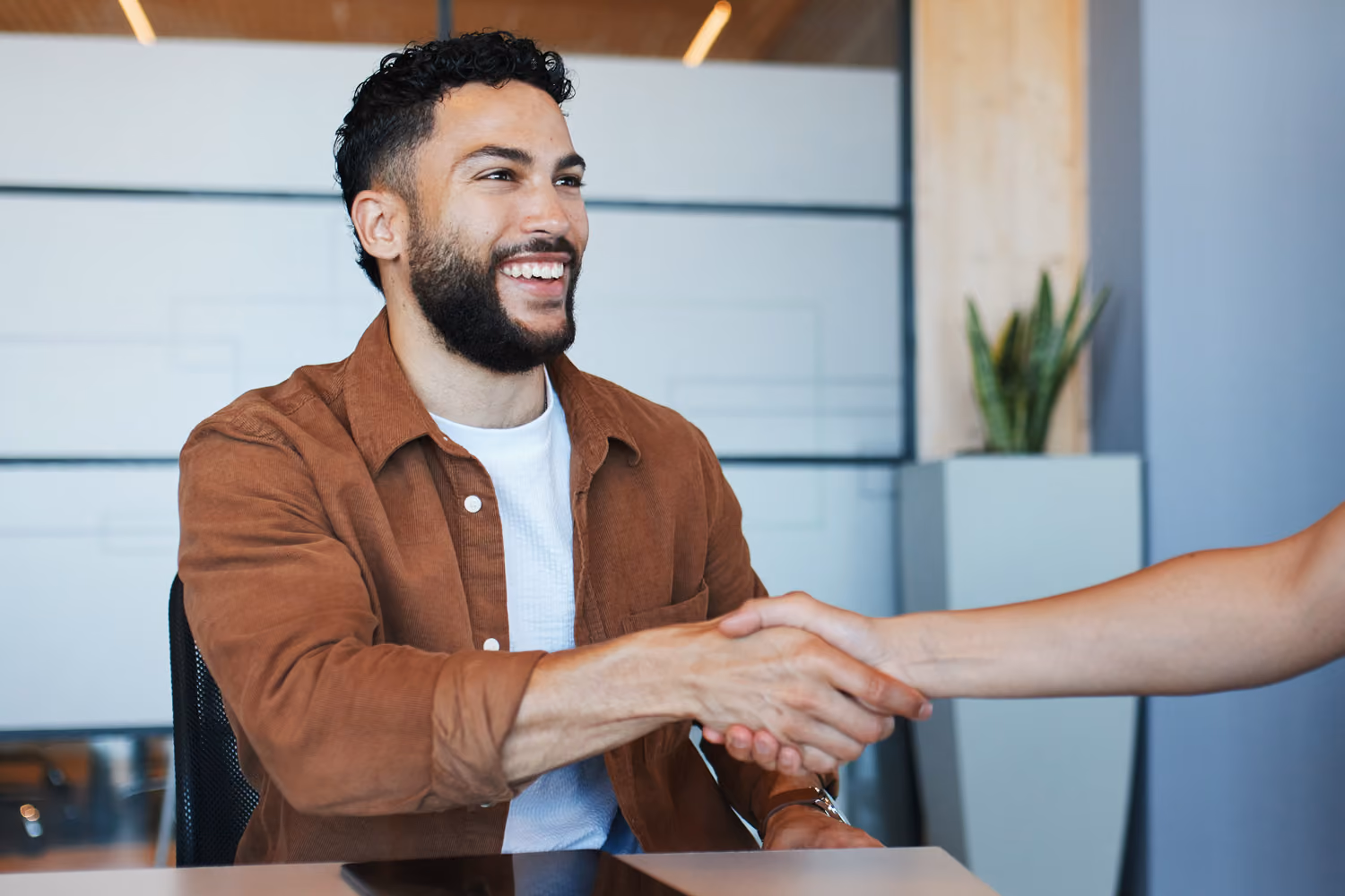 Smiling man in brown shirt shaking hands with another person in an office setting.