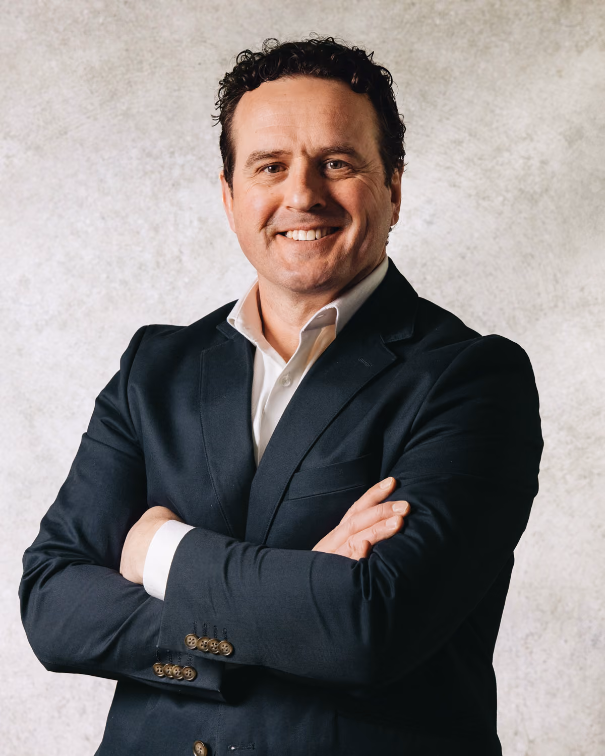 Smiling man in black suit jacket and white shirt standing with arms crossed against a light background.