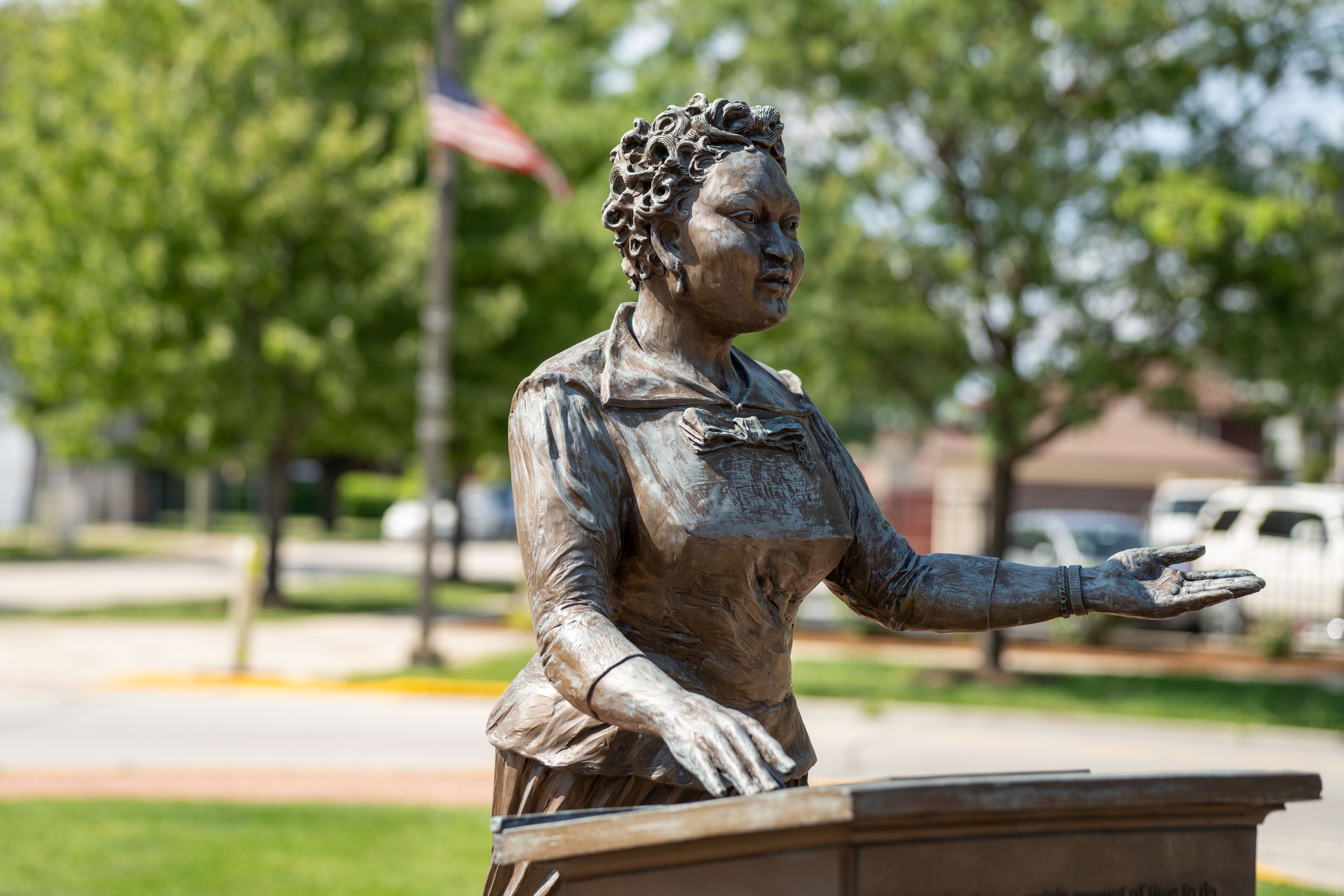 A bronze statue of a woman standing at a podium forefronts green trees and a raised American flag.