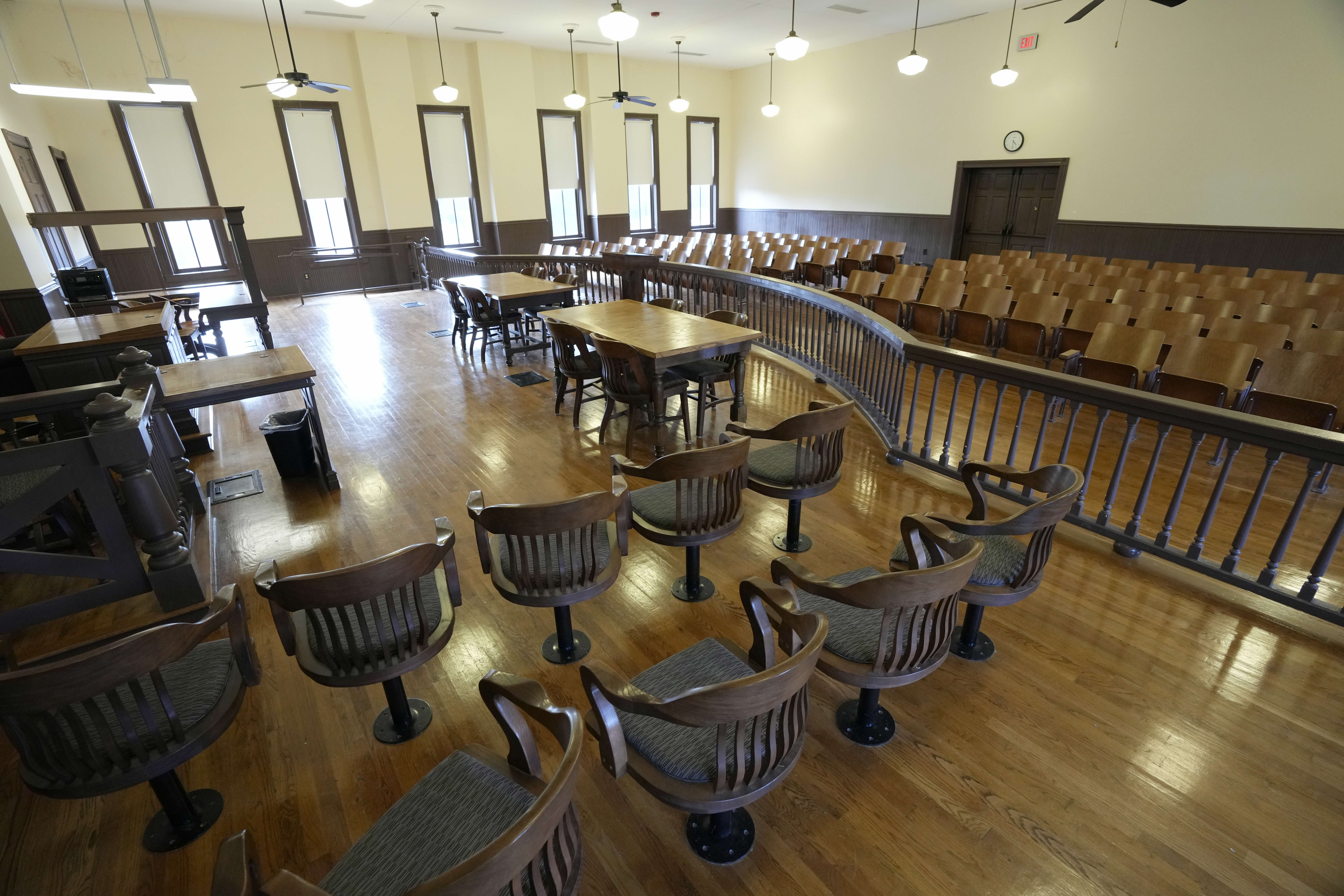 A photograph of an empty courtroom with light wooden floors and dark wooden chairs.