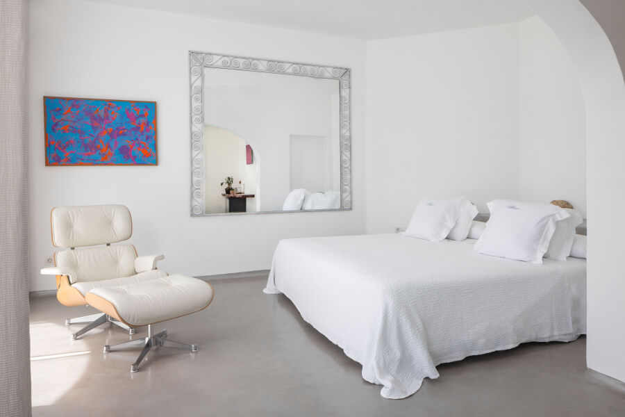 Chambre blanche de l'Hacienda Na Xamena avec grand lit, fauteuil et tableau coloré.