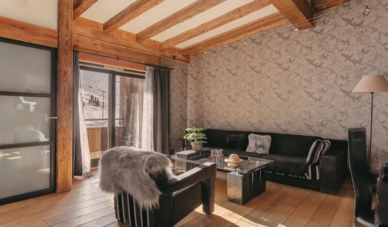 Salon de la Suite Tradition des Suites Maison Bouvier à Tignes, avec canapé sombre, poutres et porte vers le balcon.