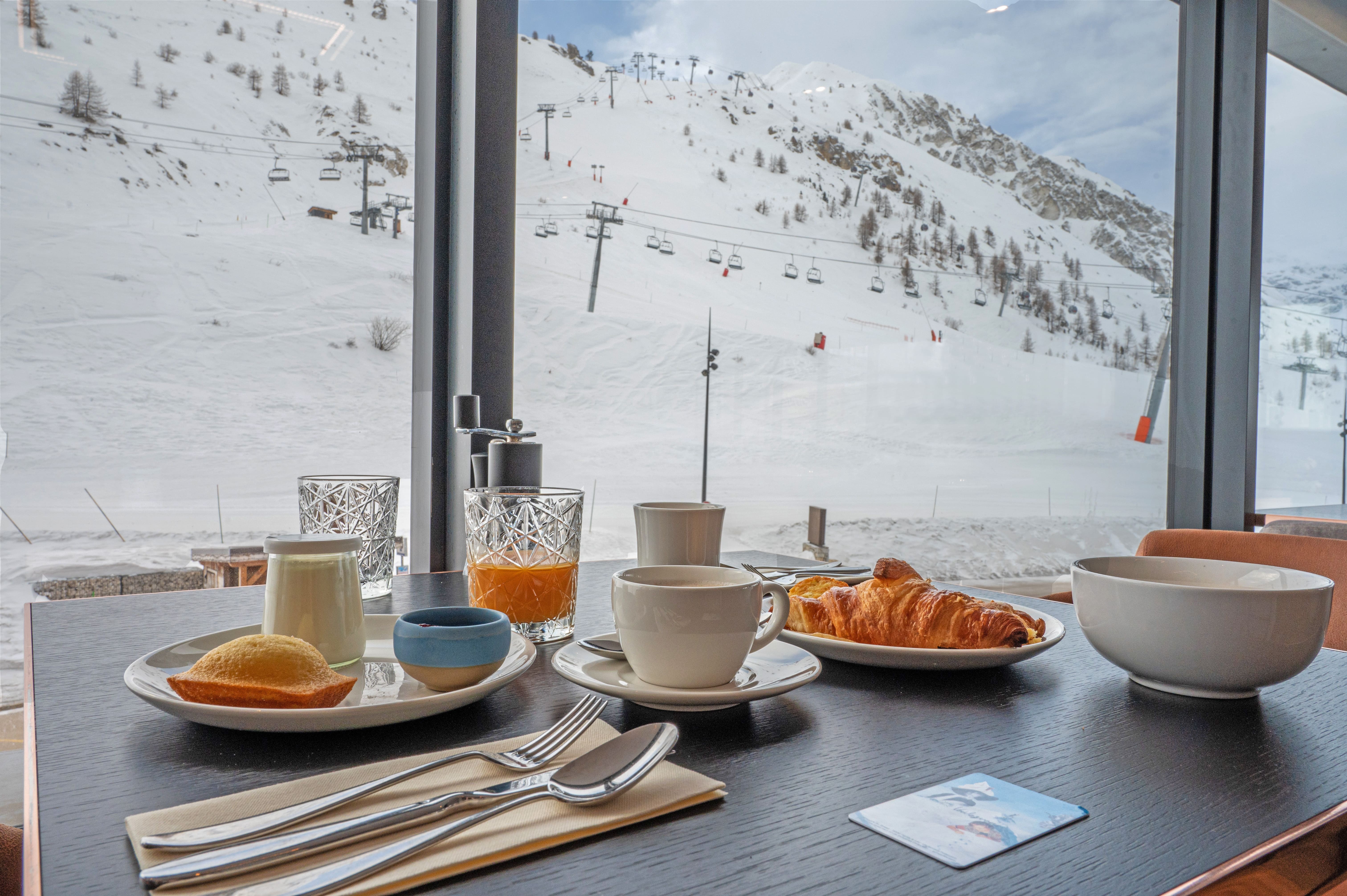 Petit-déjeuner au VoulezVous à Tignes, avec viennoiseries, tasses et jus de fruits devant les pistes enneigées.
