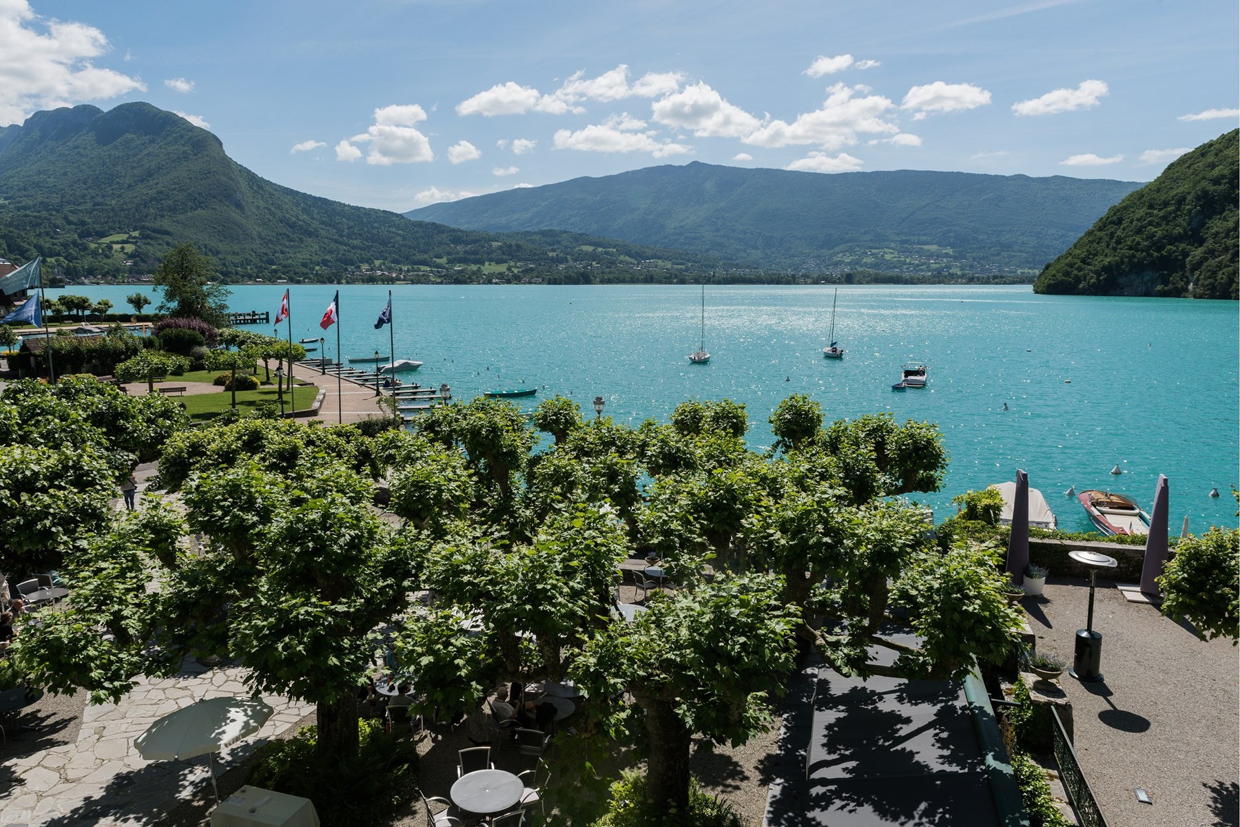 Terrasse arborée de l'Abbaye de Talloires et bateaux dans la baie, avec les montagnes au-delà du lac d'Annecy.