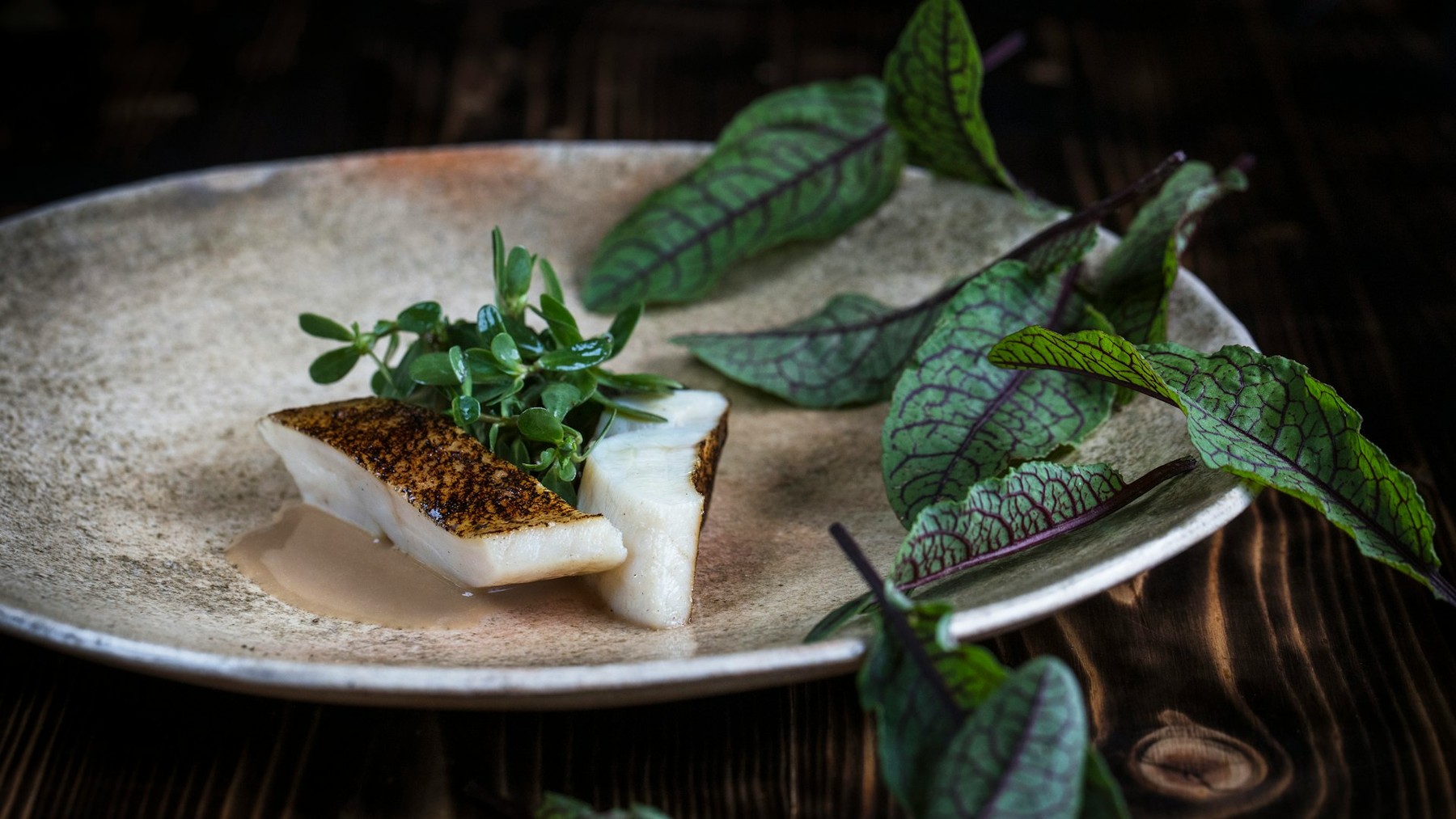 Assiette de poisson et feuilles vertes présentée par le restaurant Le Clos des Sens à Annecy-le-Vieux.