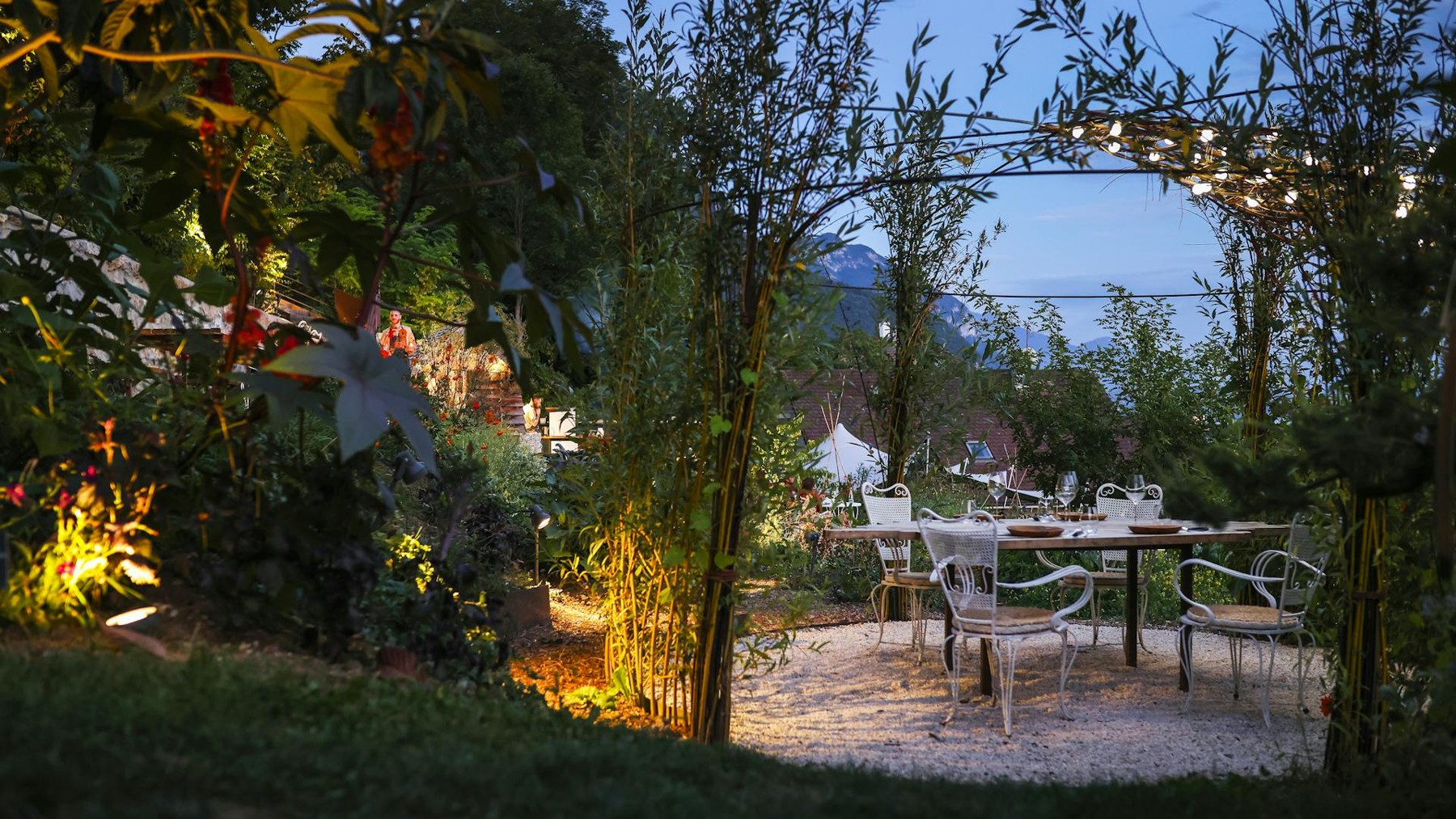 Tables et chaises sous une arche végétale éclairée dans le jardin du Clos des Sens à Annecy-le-Vieux.