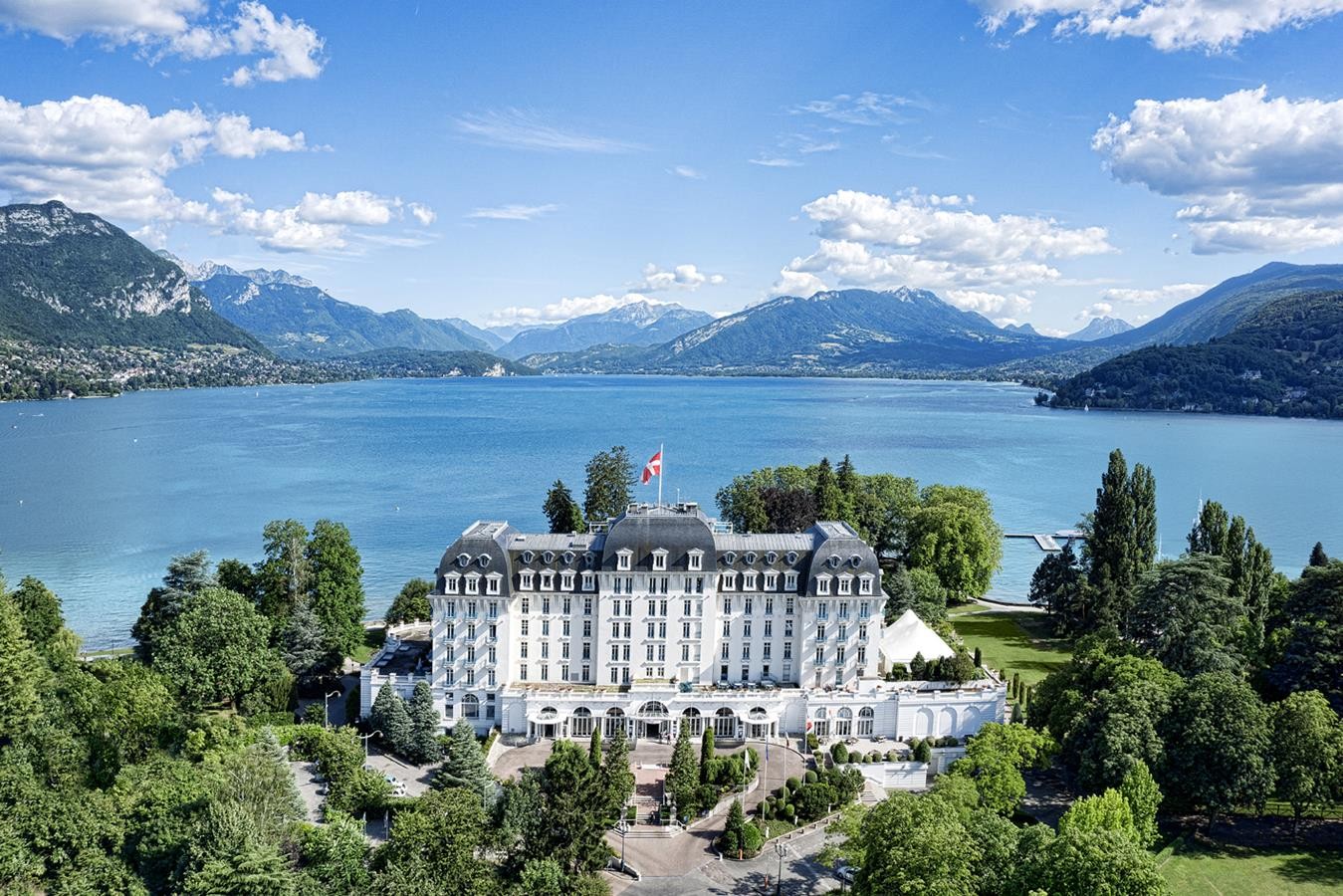Vue aérienne de la façade blanche de l'Impérial Palace, entourée de verdure devant le lac d'Annecy.