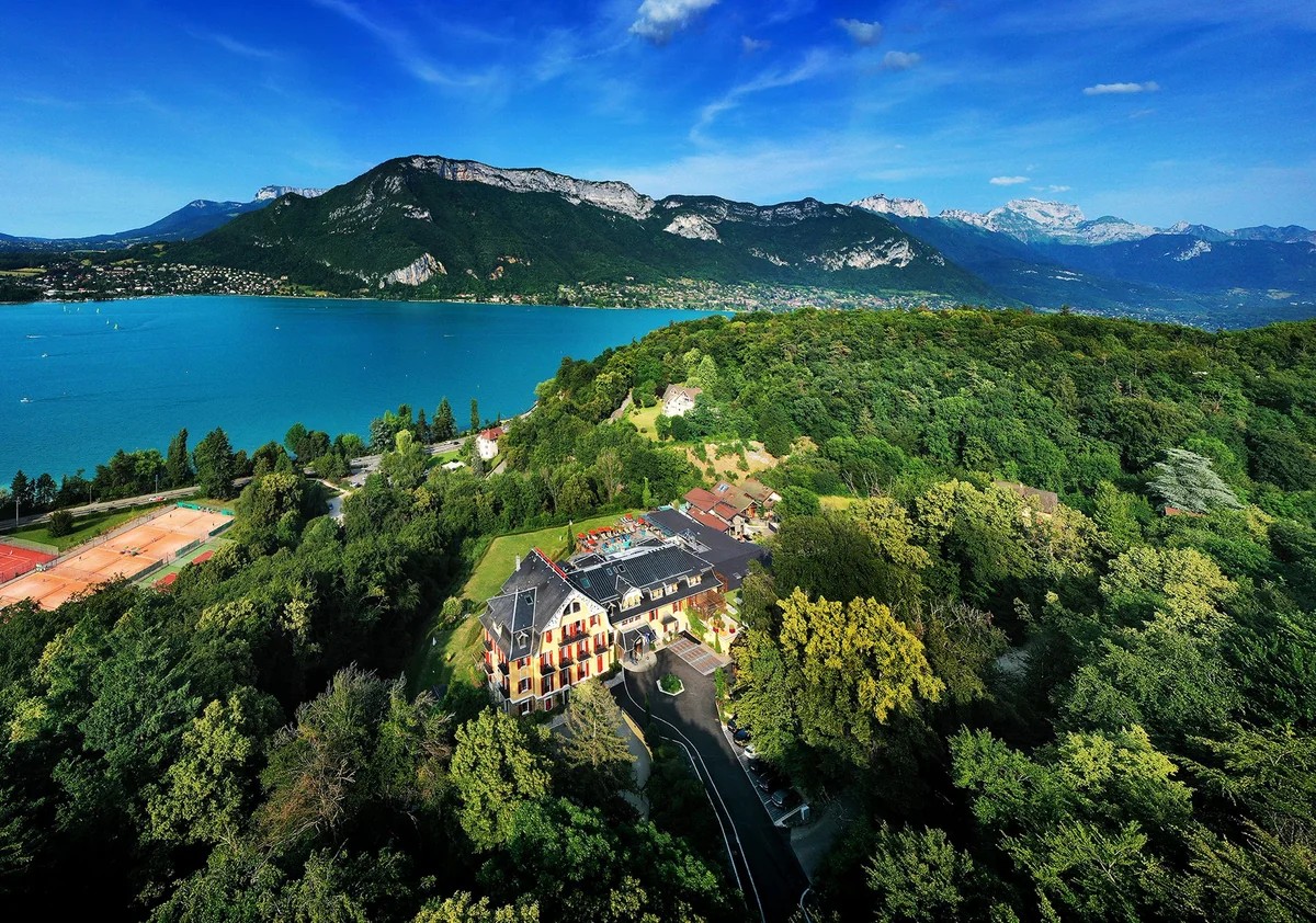 Vue aérienne des Trésoms au milieu des arbres, avec le lac d'Annecy et les montagnes en arrière-plan.