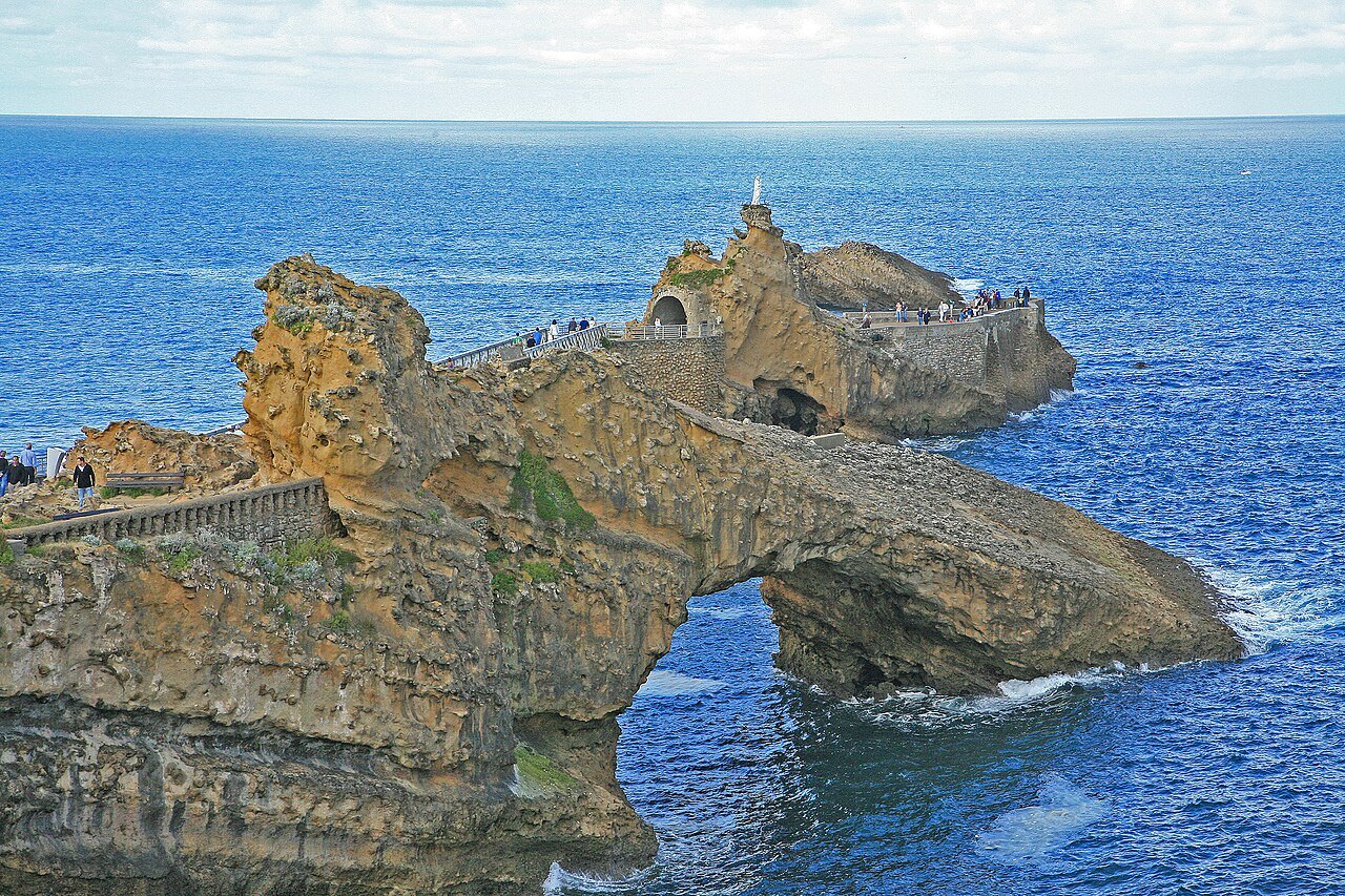 Le Rocher de la Vierge à Biarritz, sa statue blanche et sa passerelle métallique, derrière l'arche rocheuse de la Roche percée