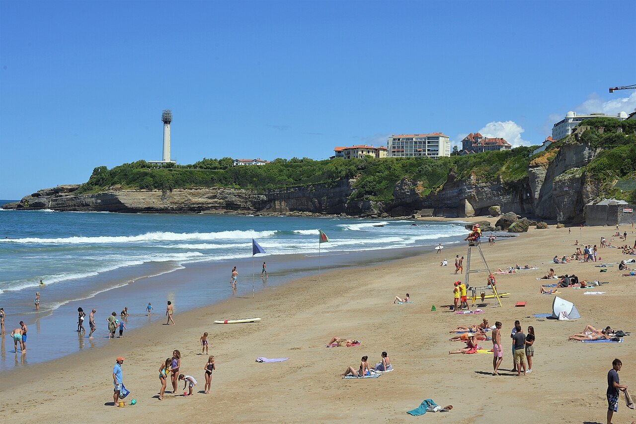 La plage du Miramar à Biarritz en été, baigneurs et poste de sauveteurs au pied de la falaise, avec le phare sur la pointe Saint-Martin