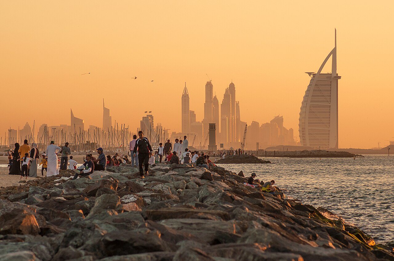 Promeneurs sur la digue de Jumeirah au coucher du soleil, avec Burj Al Arab et les tours de la Marina au loin