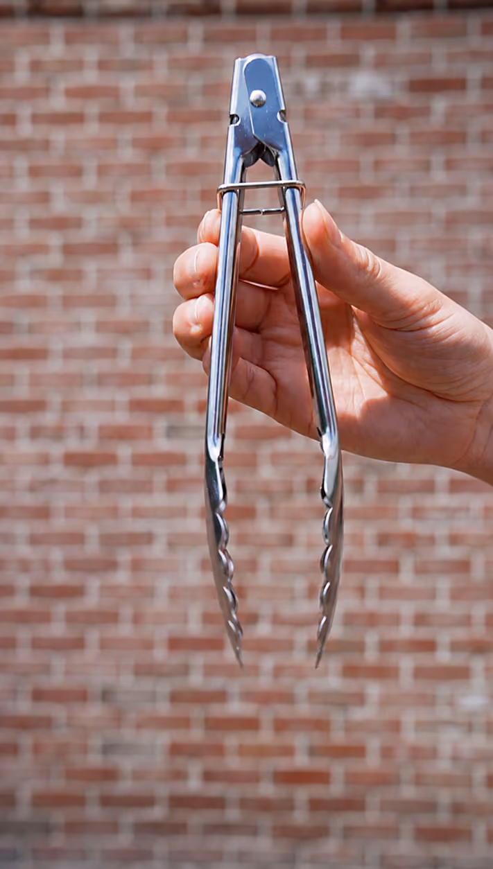 blue2purple - A hand holding stainless steel barbecue tongs in front of a red brick wall