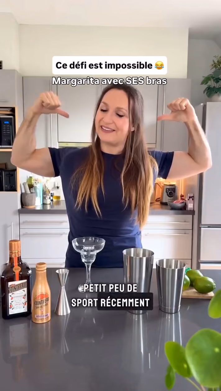 blue2purple - A woman flexing her arms behind a kitchen counter set with cocktail shakers and a glass