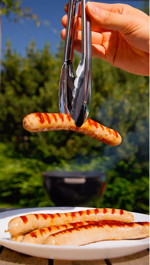 blue2purple - A hand using tongs to lift grilled sausages above a plate in a garden