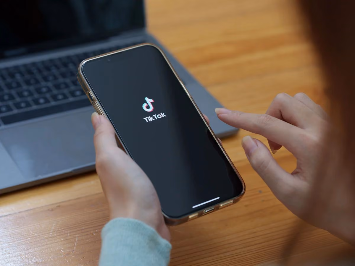 blue2purple - Hand holding a phone with the TikTok logo in front of a laptop