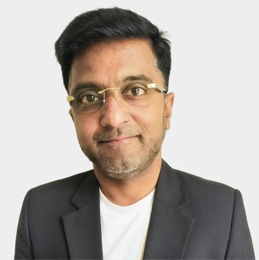 Man wearing glasses, a black blazer, and a white shirt smiling slightly on a light background.