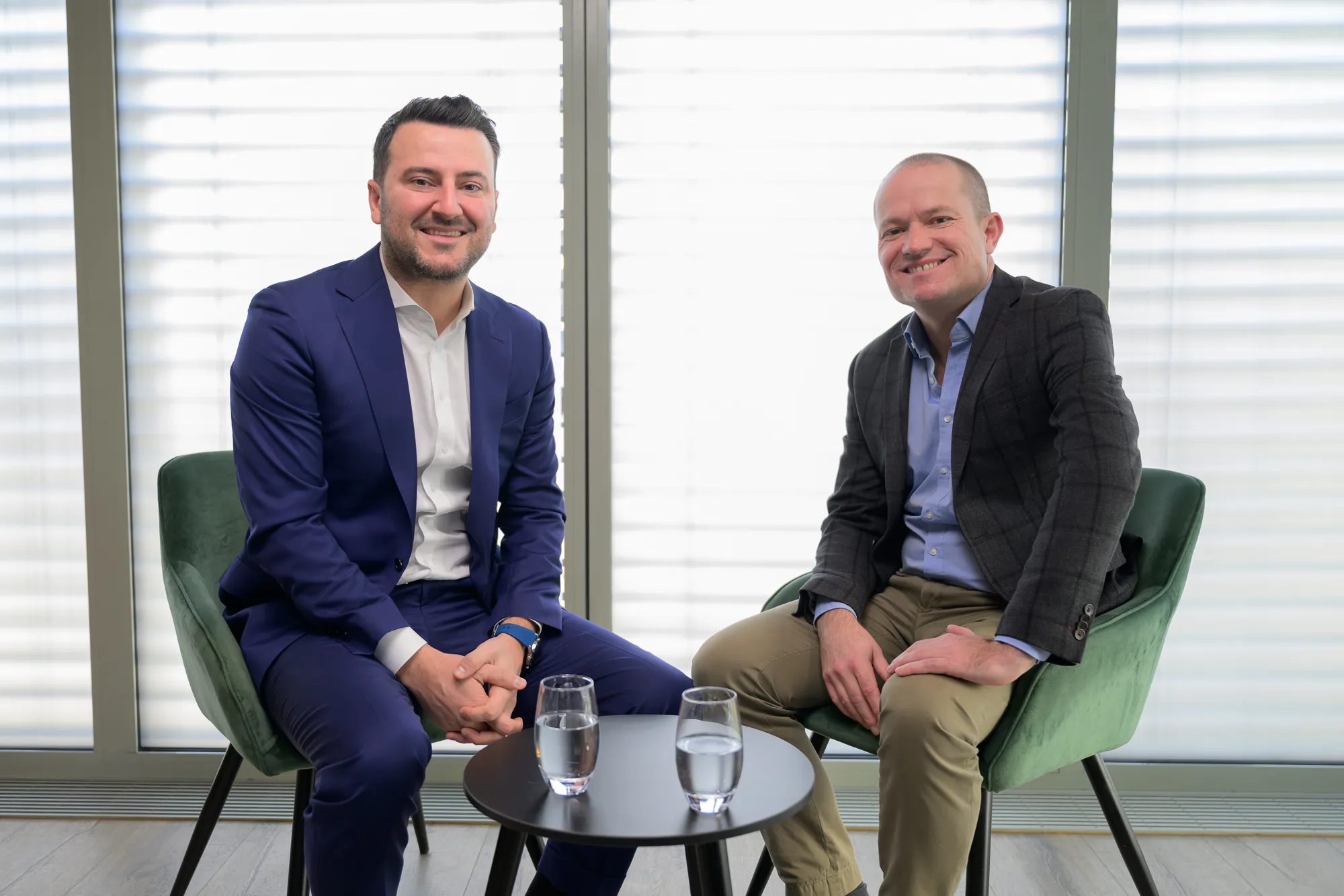 Two men in business attire sitting on green chairs by a small round table with water glasses.