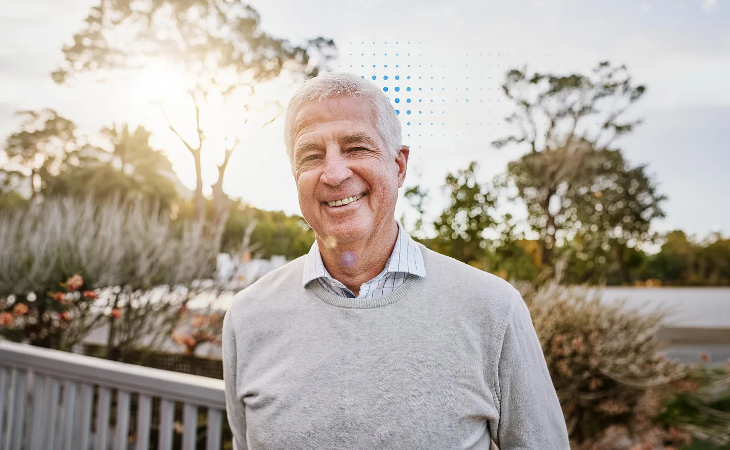 Smiling older man with gray hair wearing a light gray sweater and collared shirt, standing outdoors with trees and soft sunlight in the background.
