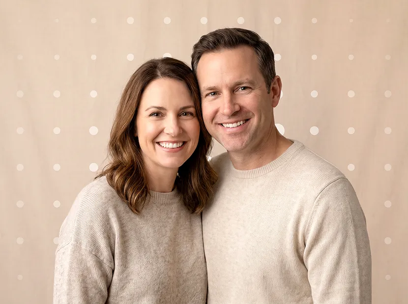 Smiling man and woman wearing beige sweaters standing close against a beige background with dots.