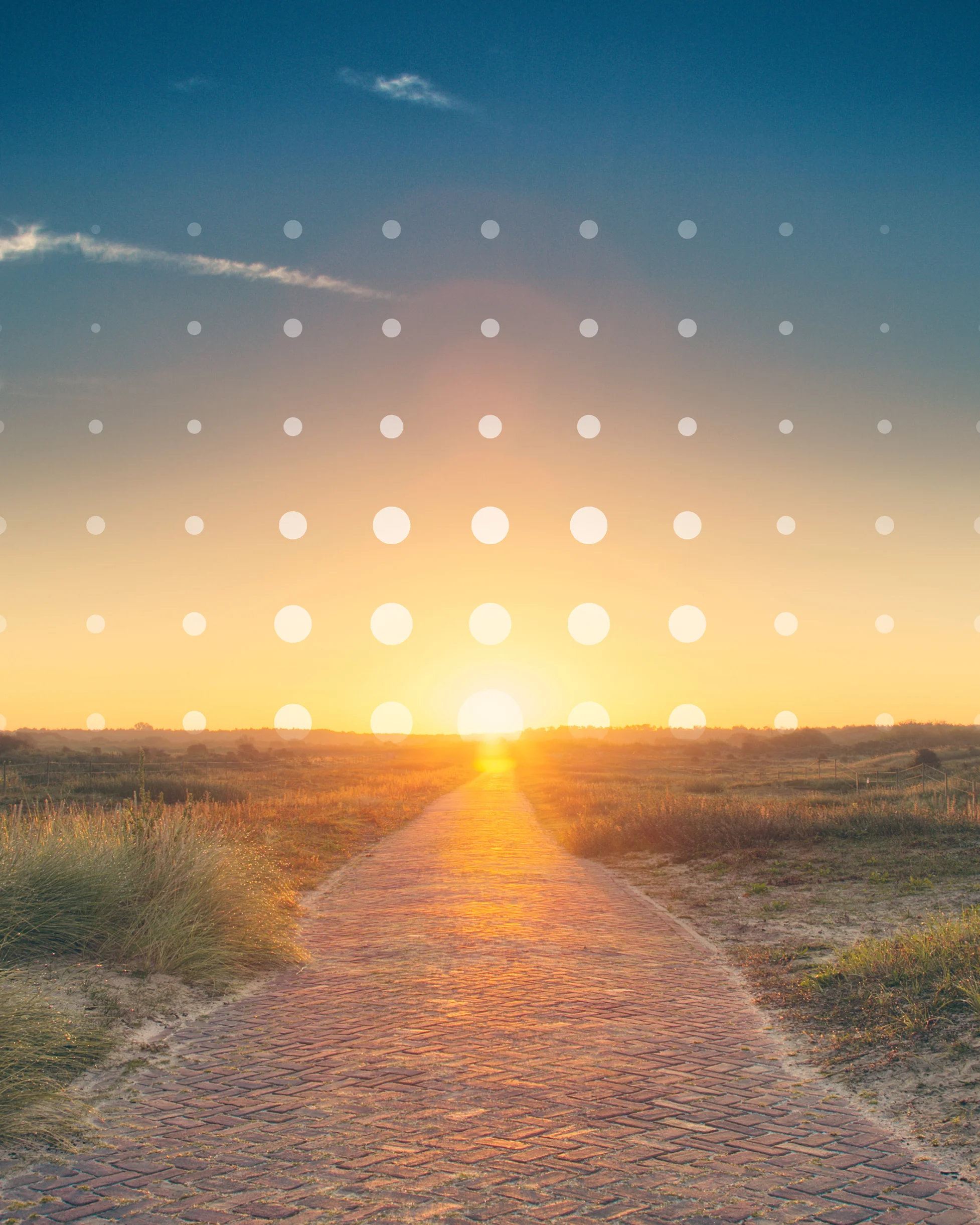 Sunset over a brick path through fields with grass under a partly cloudy blue sky.