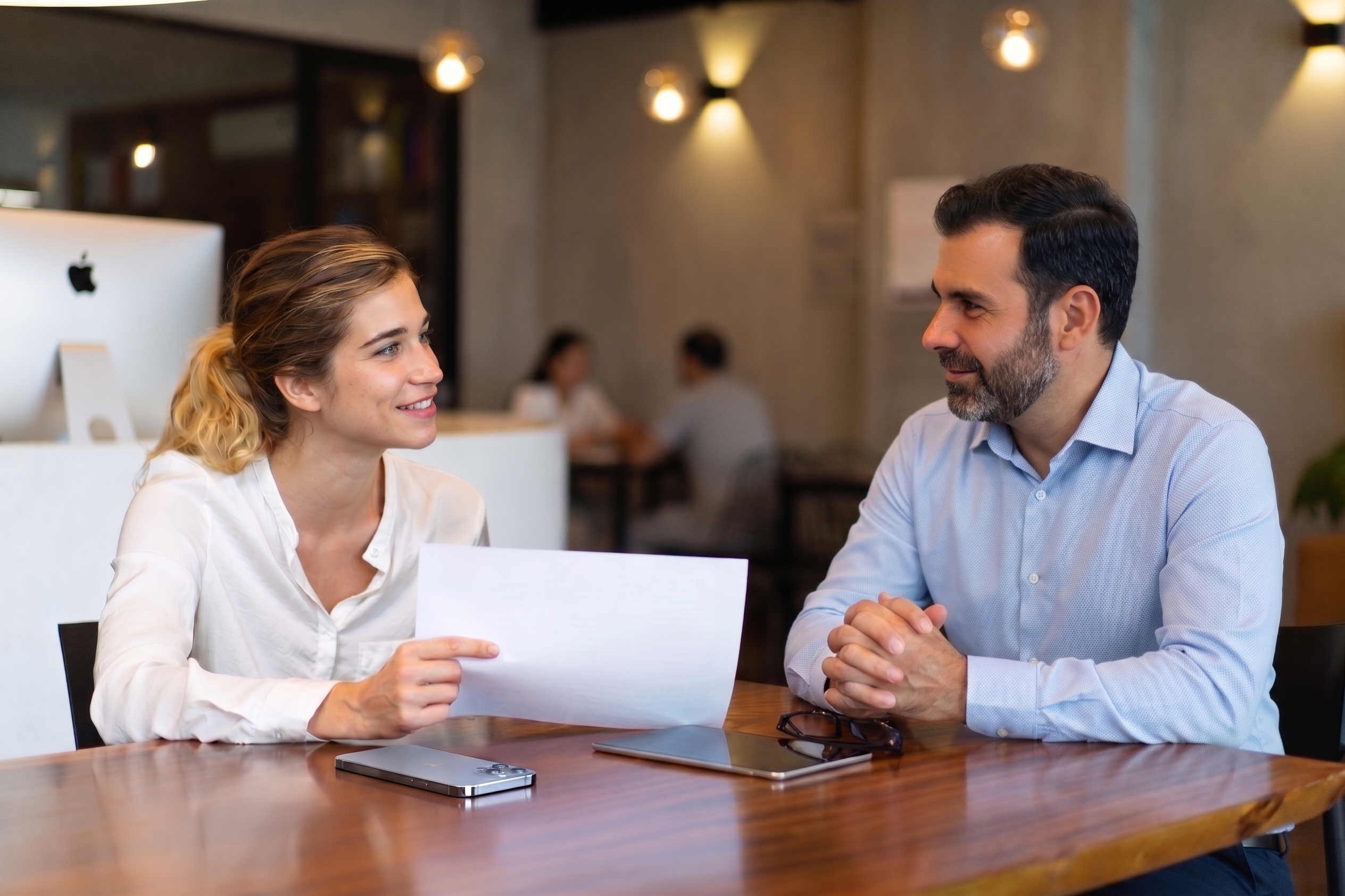 Woman showing a document to a man while they sit at a wooden table in an office setting.