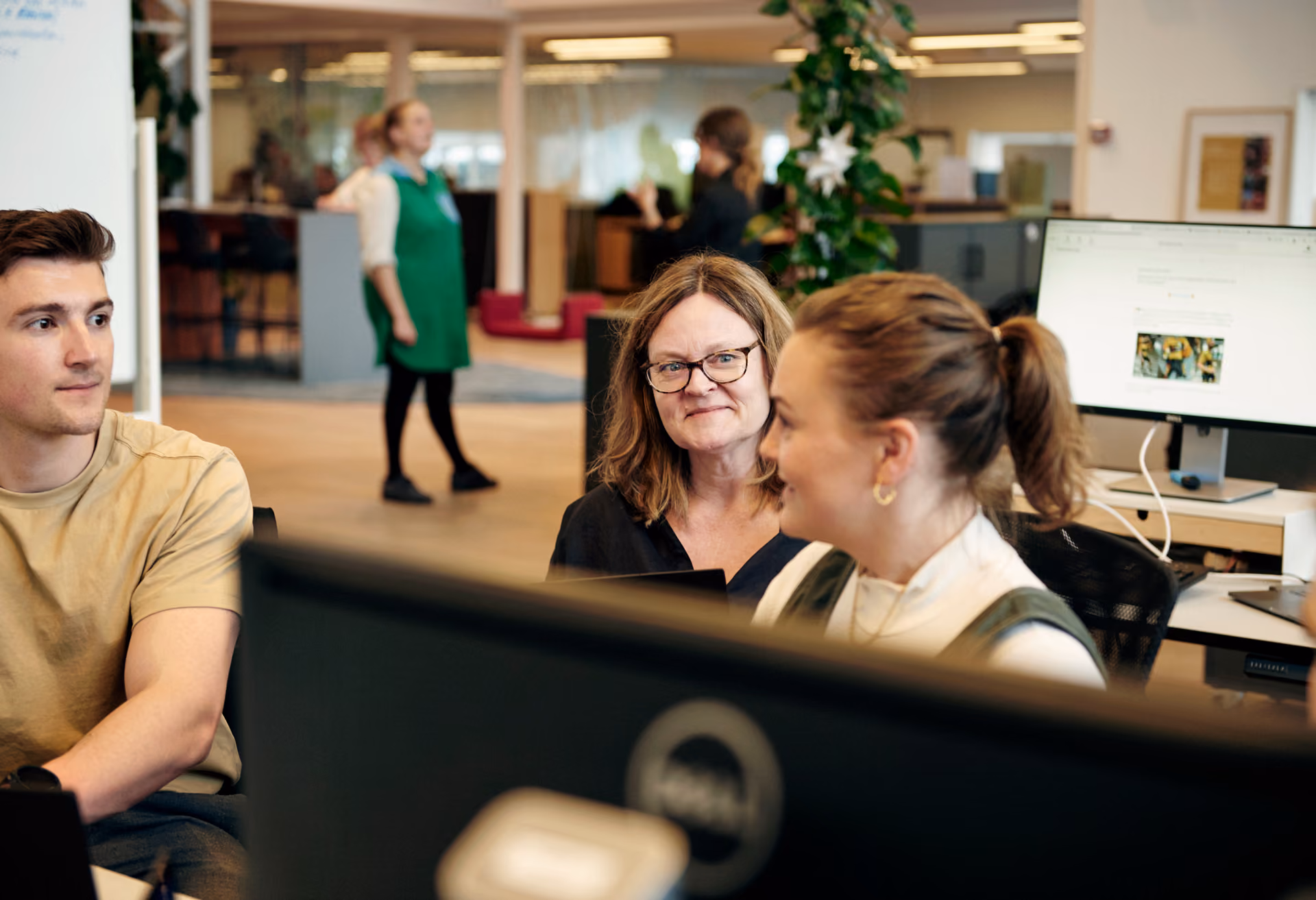 Tre personer sitter och pratar i ett öppet kontorslandskap med datorskärmar i förgrunden.