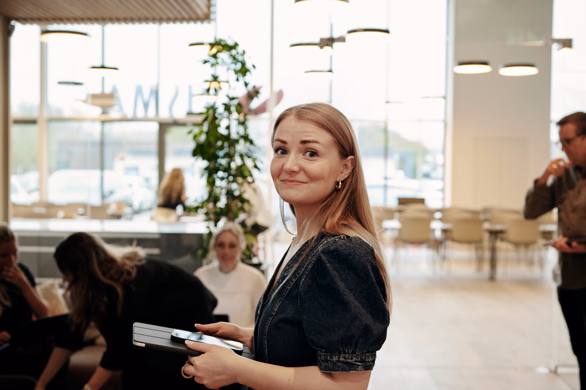 Kvinna med långt blont hår håller en surfplatta och mobil inomhus med personer i bakgrunden.