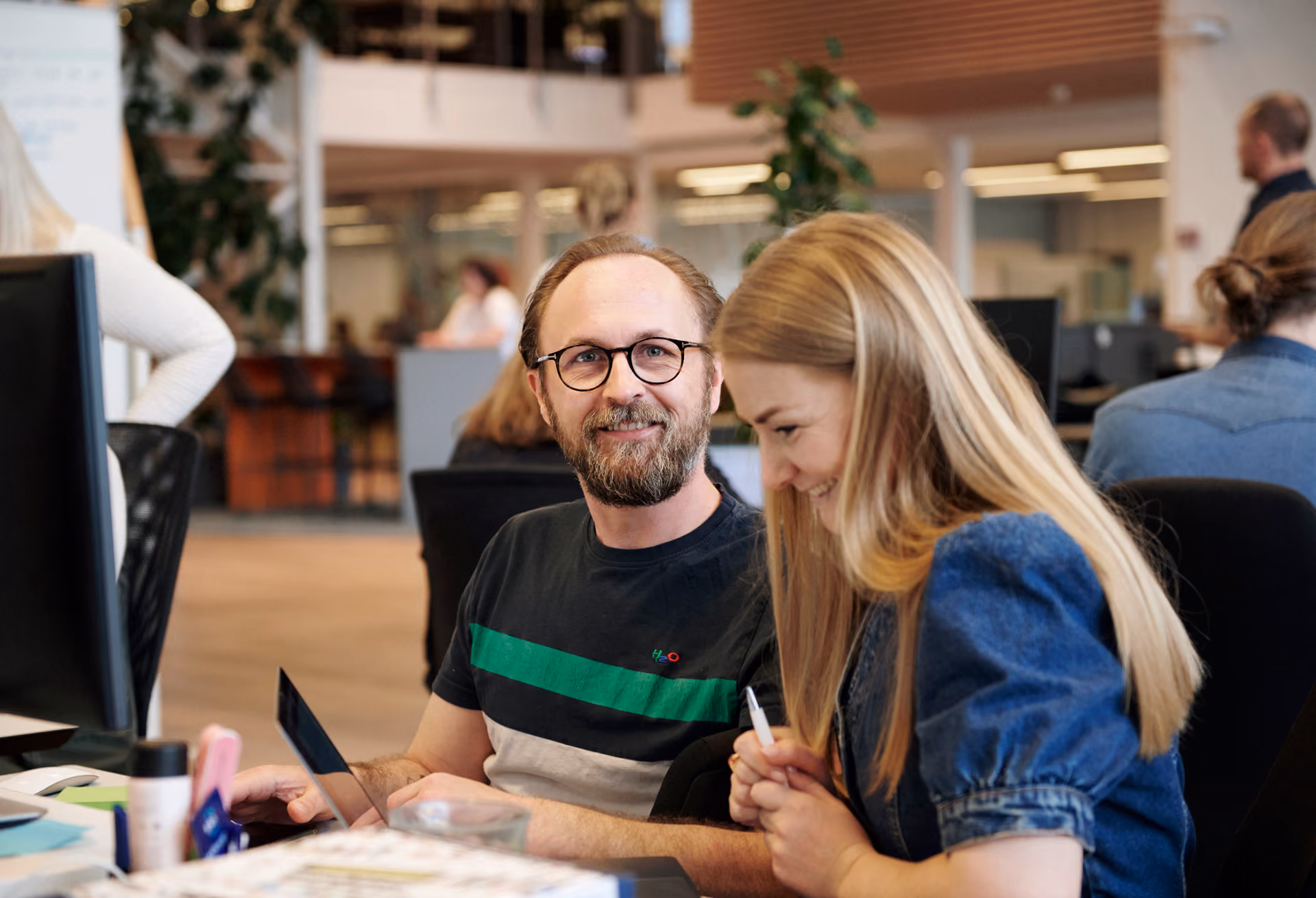 En man och en kvinna ler och arbetar tillsammans på ett kontor med datorer i bakgrunden.