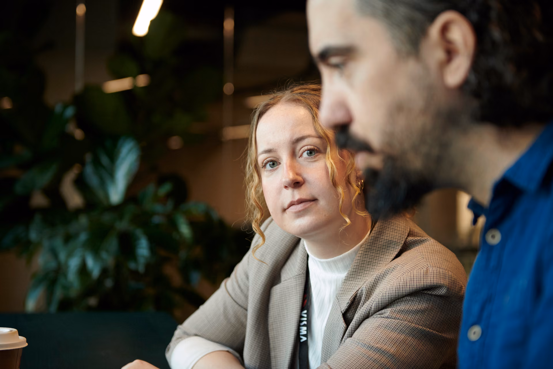 Kvinna i kavaj ser på en man medan de sitter inomhus med växter i bakgrunden.