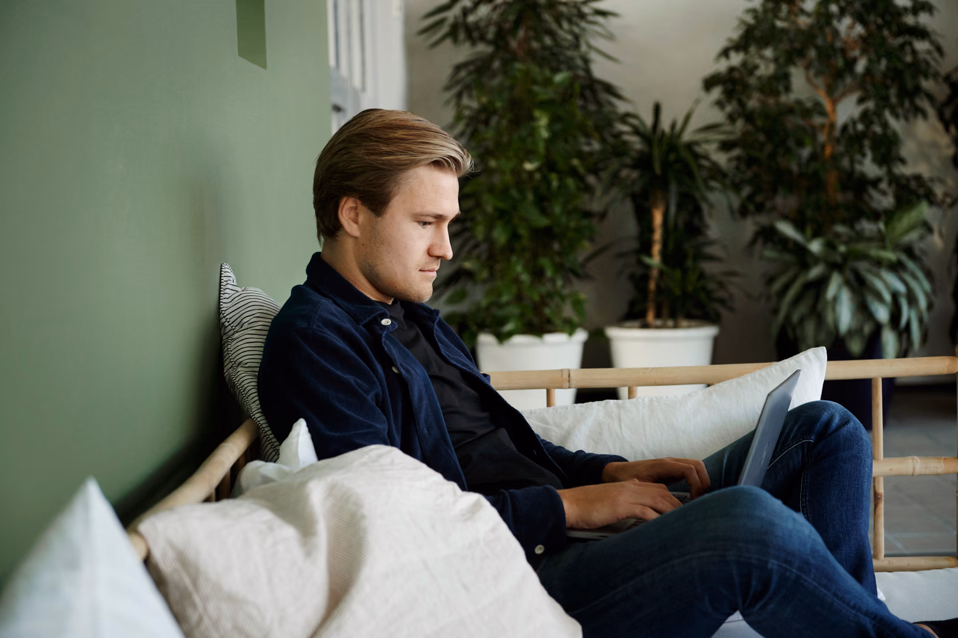 Ung man med blont hår sitter avslappnad i en soffa och arbetar på sin laptop, med krukväxter och en grön vägg i bakgrunden.