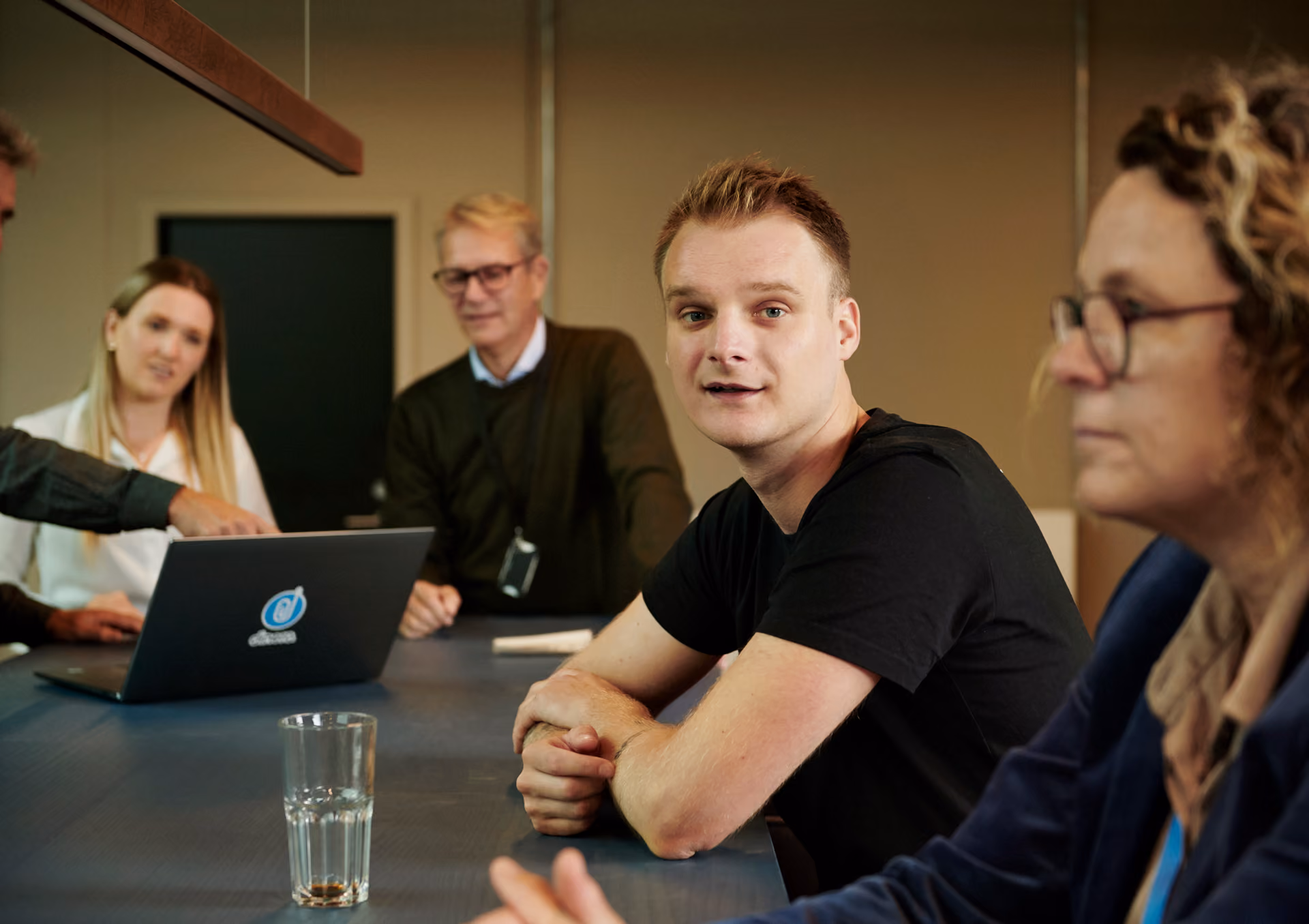 En grupp kollegor sitter runt ett mötesbord med en bärbar dator och samtalar, med en ung man i svart t-shirt i fokus.