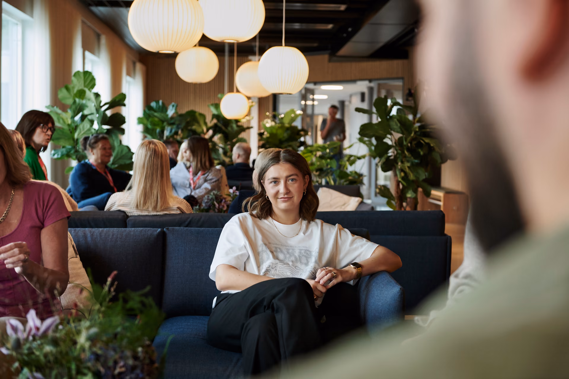 Kvinna i vit t-shirt sitter avslappnat i en mörkblå soffa i ett ljust kontorslandskap med runda hänglampor, gröna växter och kollegor i bakgrunden.