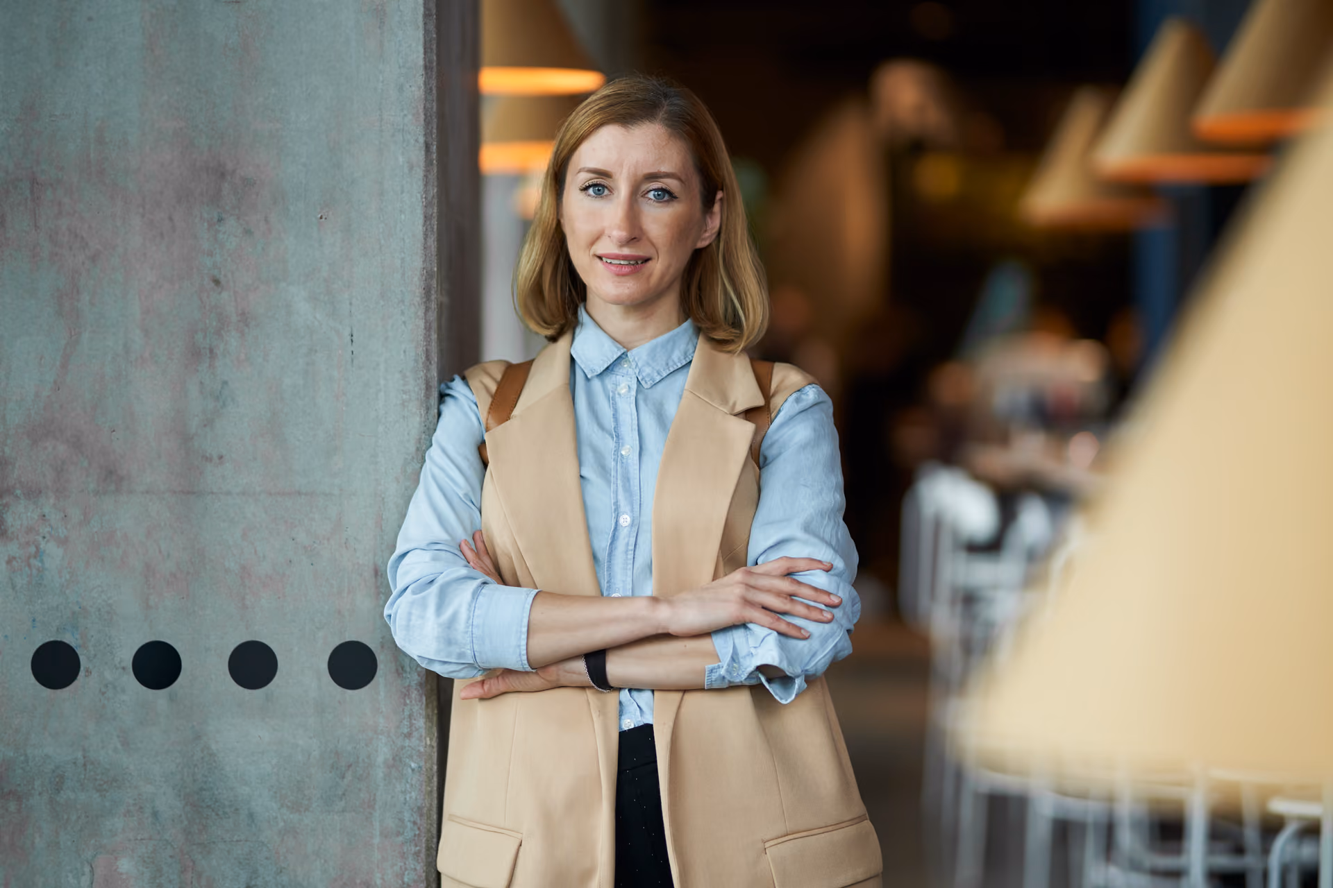 Kvinna i ljusblå skjorta och beige väst står med armarna i kors lutad mot en betongpelare i en restaurangmiljö med hängande lampor i bakgrunden.