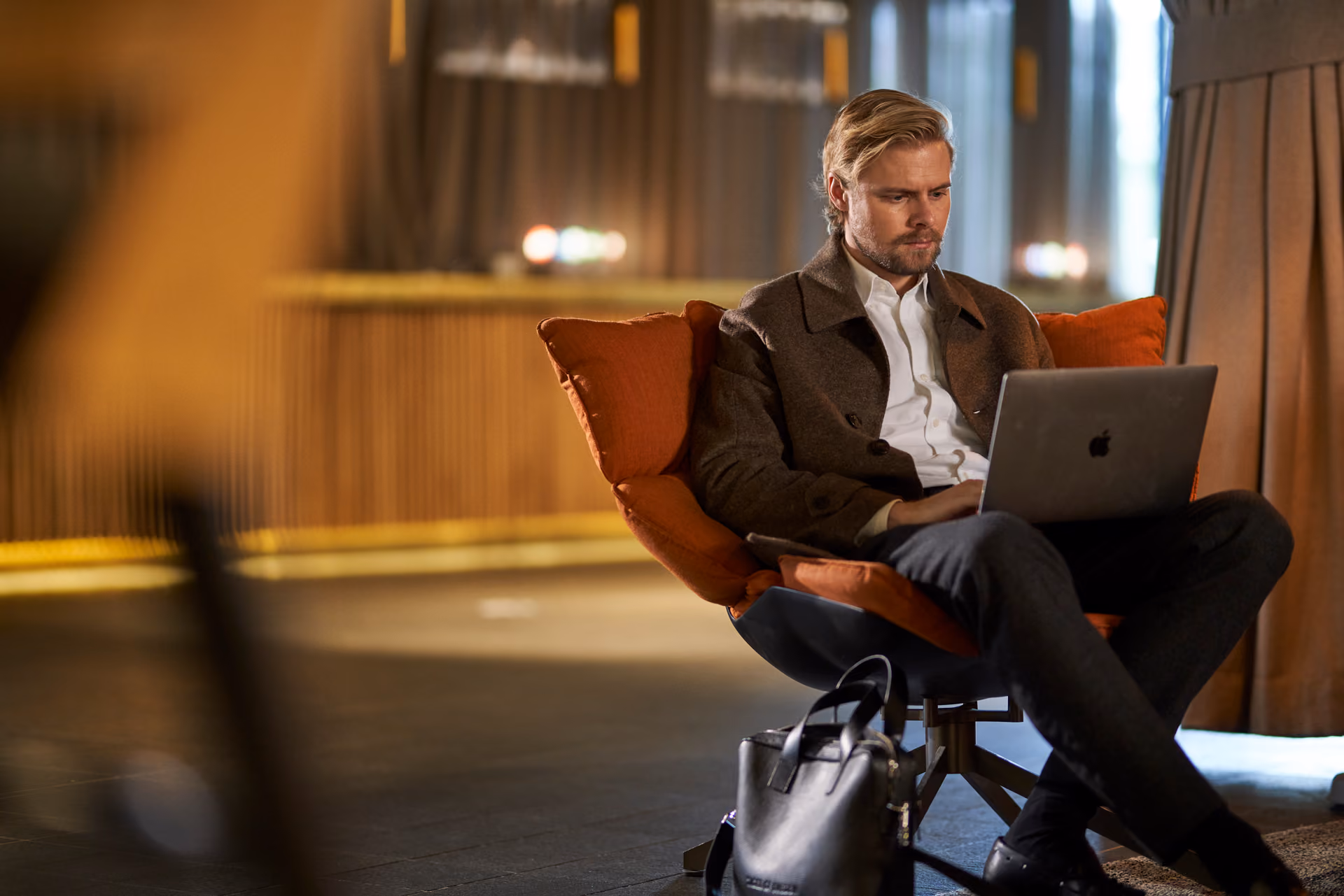 Blond man i brun rock sitter i en fåtölj och arbetar på en bärbar dator i en stämningsfull lobby med dämpad belysning.