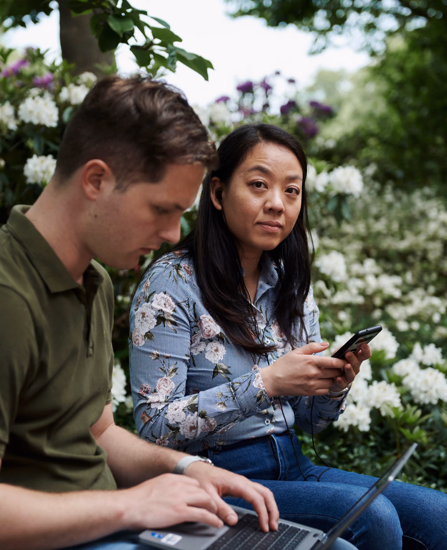 Man arbetar vid en bärbar dator medan en kvinna i blommig skjorta håller en mobiltelefon, sittande utomhus framför blommande buskar och grönska.