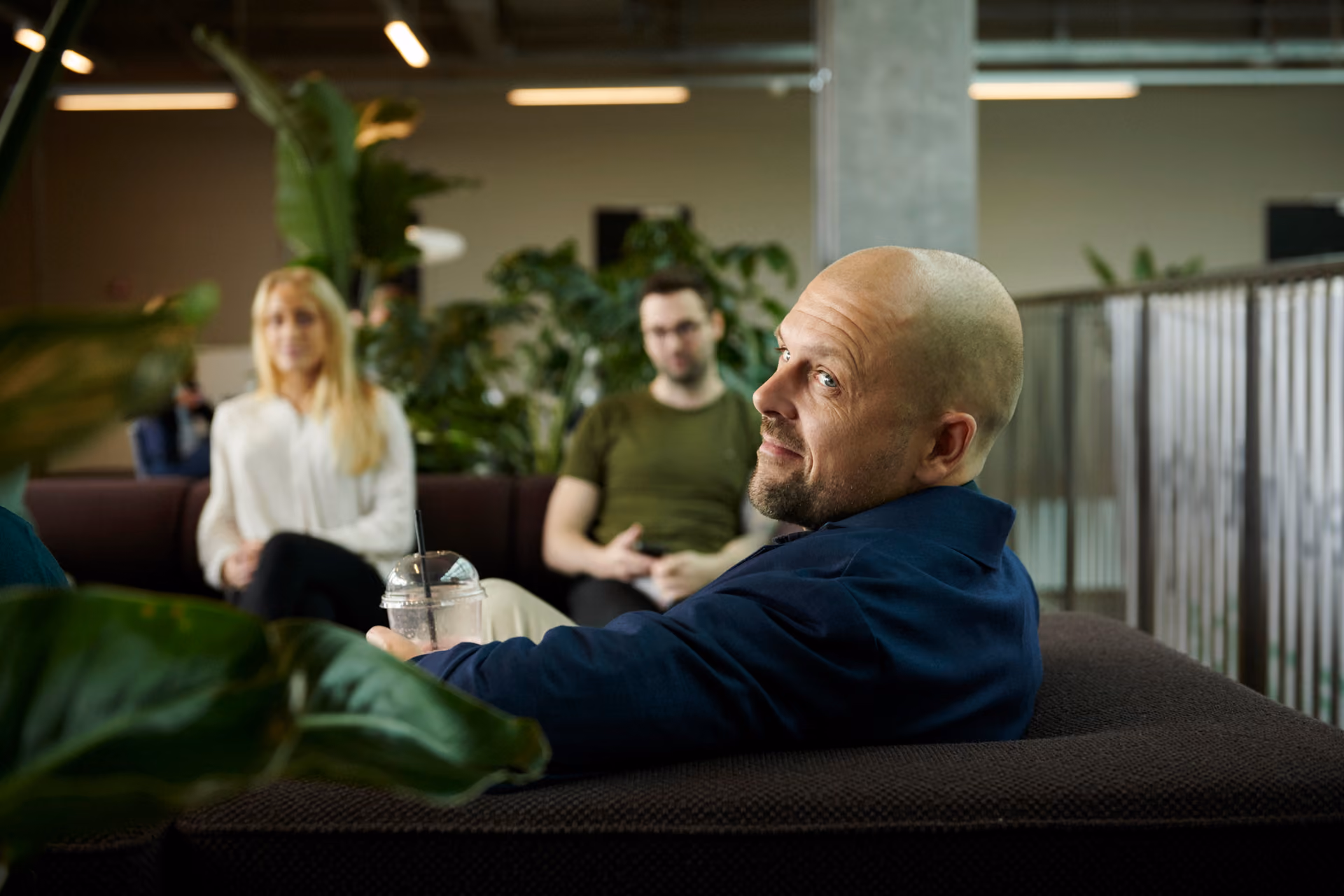 Skallig man i mörkblå kavaj sitter i en soffa med en iskaffe i handen, med två personer och gröna växter i bakgrunden.
