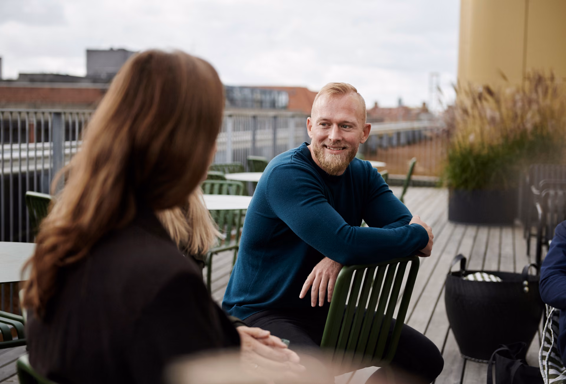 Leende man med rött skägg i mörkblå tröja sitter på en grön stol på en takterrass och samtalar med en kvinna, med hustak och prydnadsgräs i bakgrunden.