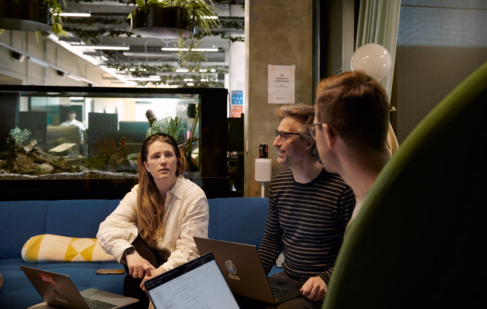 Kvinna i vit skjorta sitter i en blå soffa och samtalar med två män på ett kontor, med ett akvarium och gröna hängväxter i bakgrunden.