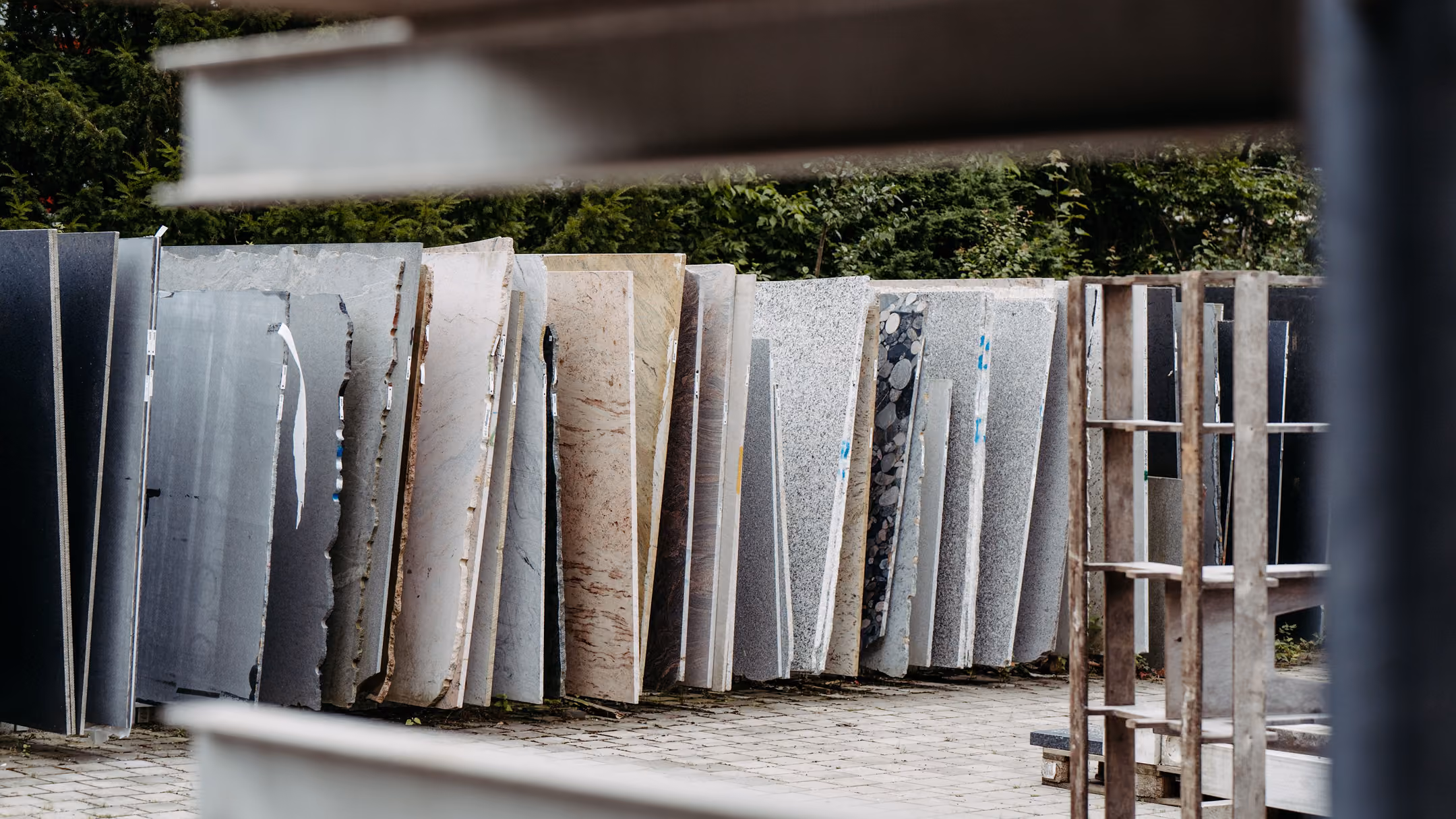 Mehrere große Steinplatten in verschiedenen Farben und Mustern stehen auf einem gepflasterten Boden im Freien, teilweise von Metallrahmen eingerahmt, mit grünen Bäumen im Hintergrund.