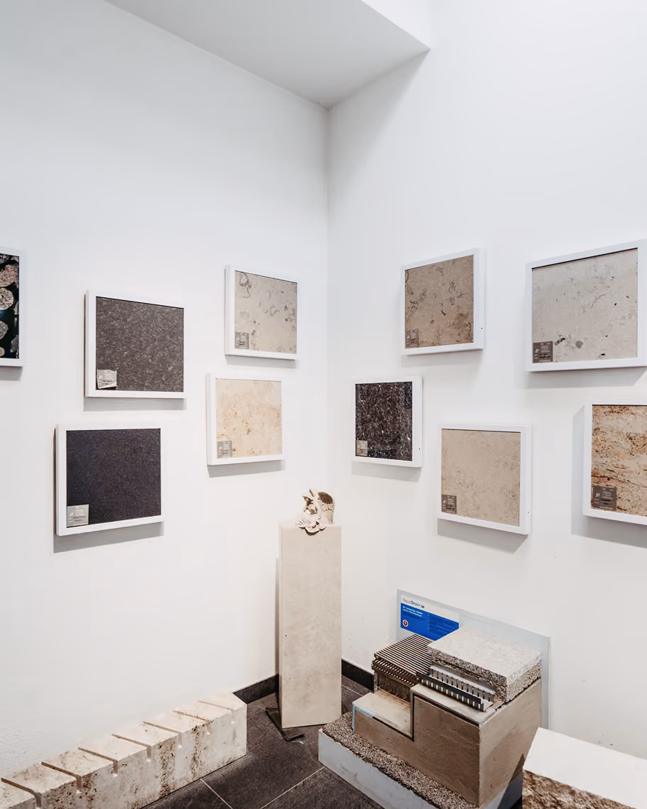 Ausstellung mit Wandtafeln aus verschiedenen Gesteins- und Steinproben in einem weißen Raum, darunter Kernbohrproben und Querschnitte, angeordnet in weißen Rahmen und auf Sockeln.
