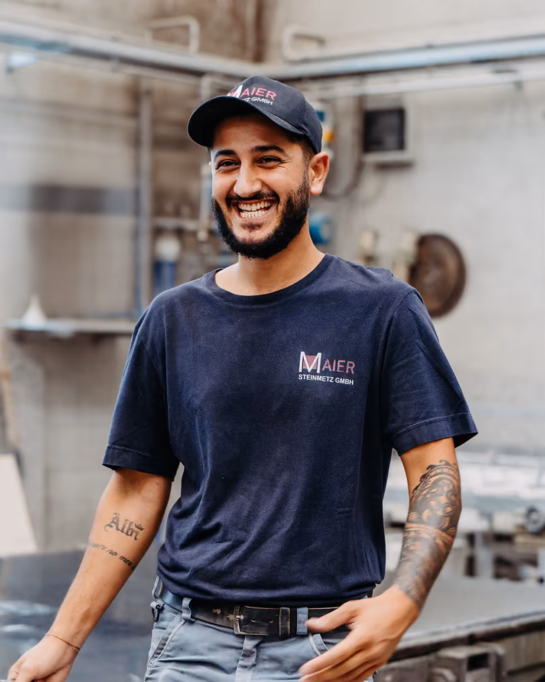 Lächelnder Mann mit kurzem Bart und Tattoos auf seinen Armen trägt eine schwarze Kappe und ein schwarzes T-Shirt mit dem Aufdruck 'Maier Steinmetz GmbH' in einer Werkstattumgebung.