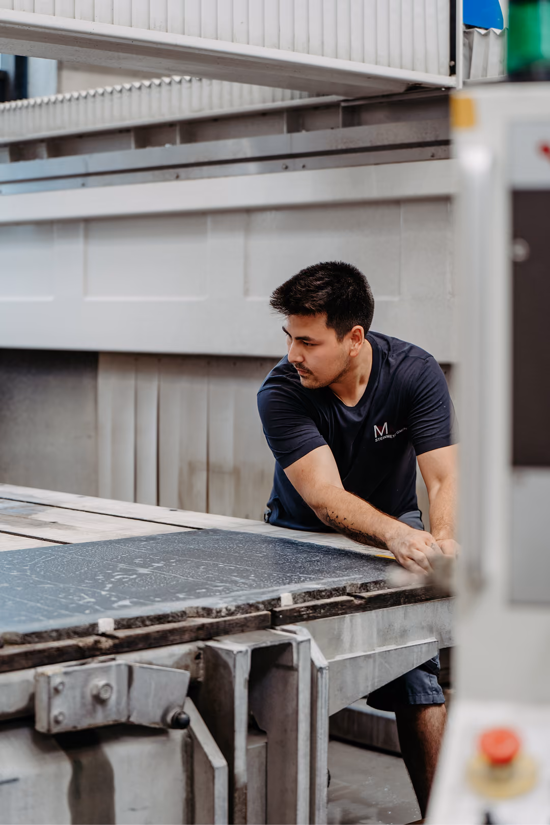 Ein Arbeiter in schwarzem T-Shirt kontrolliert eine große Steinplatte auf einem industriellen Fließband in einer Fabrikhalle.