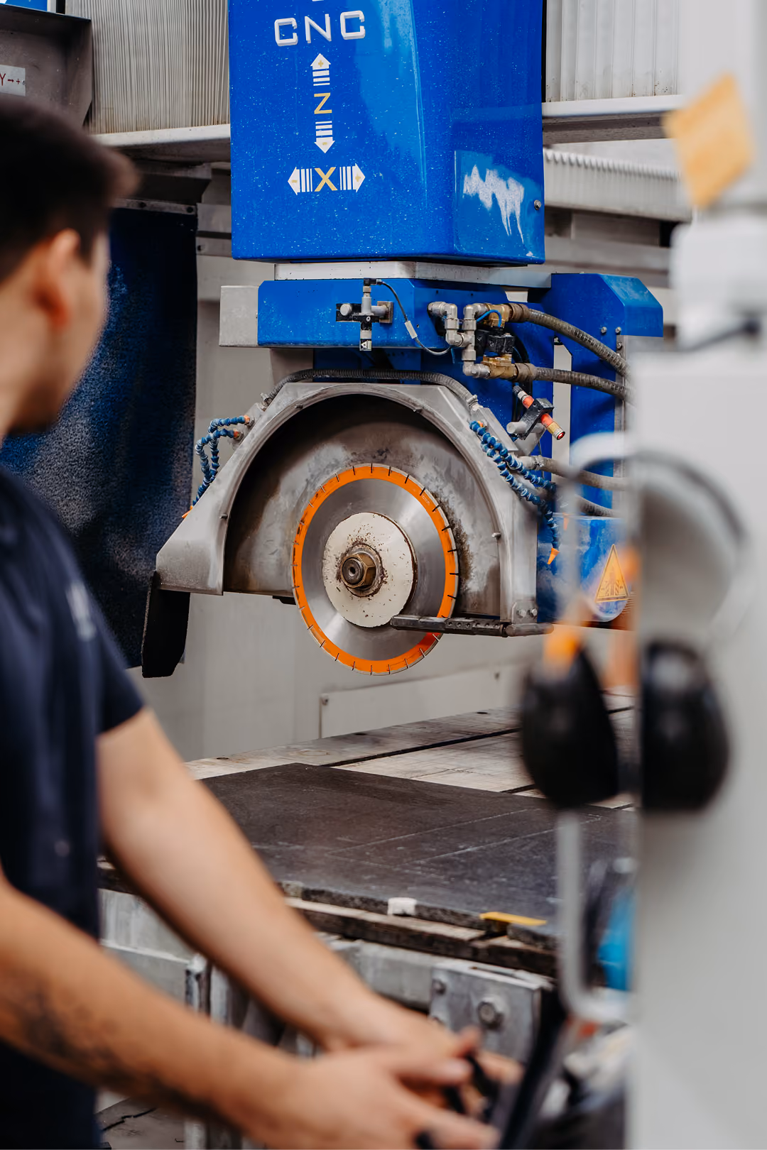 Ein Arbeiter bedient eine CNC-Maschine mit einem großen, orange markierten Sägeblatt in einer Fertigungsumgebung.