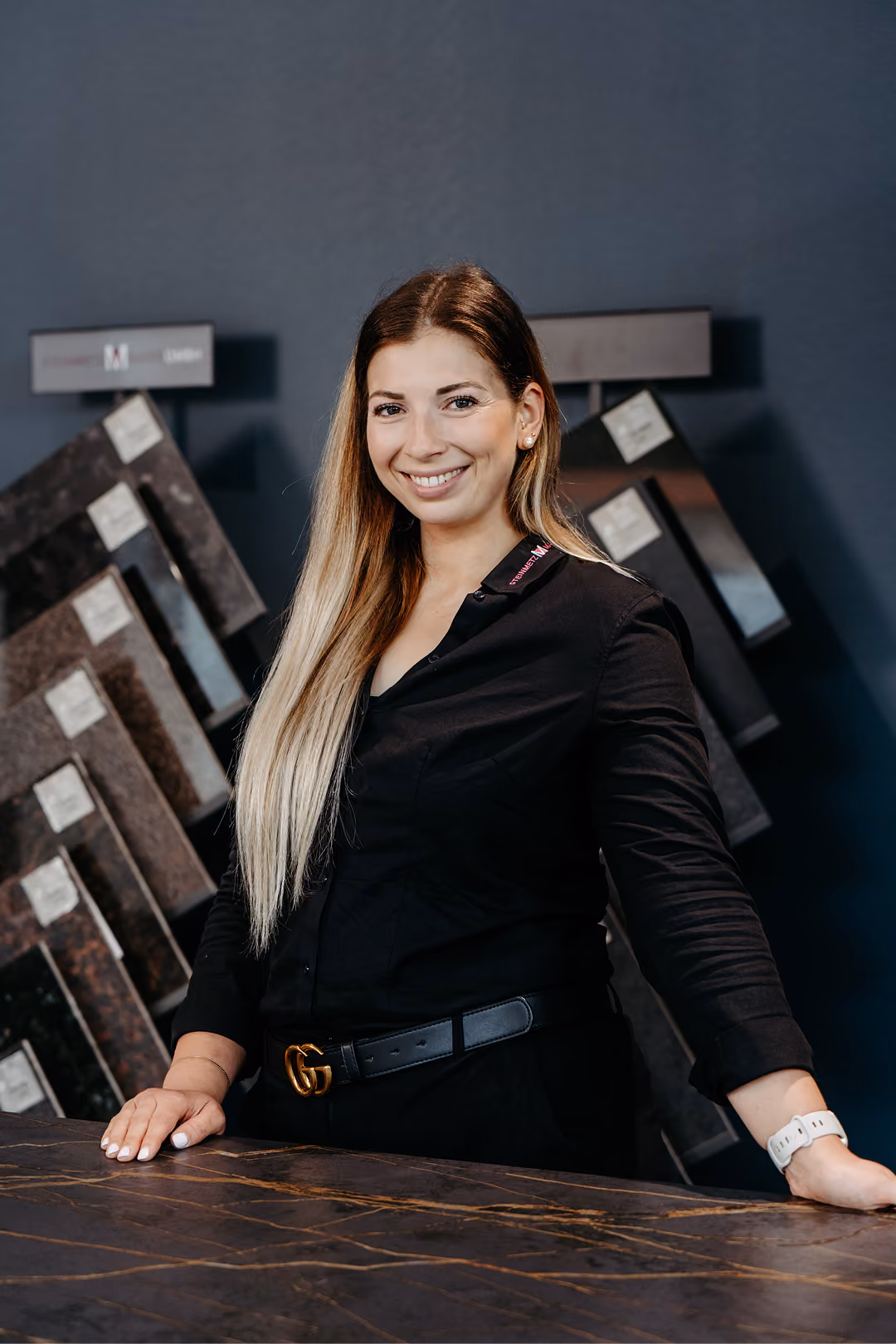 Junge Frau mit langen blonden Haaren und schwarzem Hemd steht lächelnd hinter einem Marmortisch in einem Ausstellungsraum mit Fliesenmustern an der Wand.